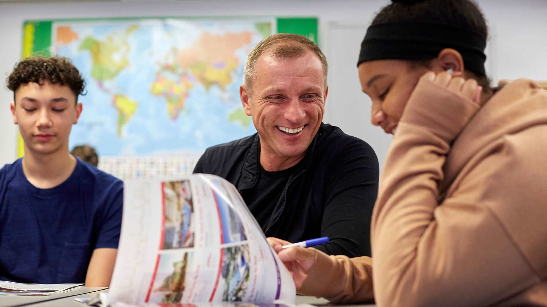 a teacher is laughing while he sits in between two of his students who are also laughing inside the classroom