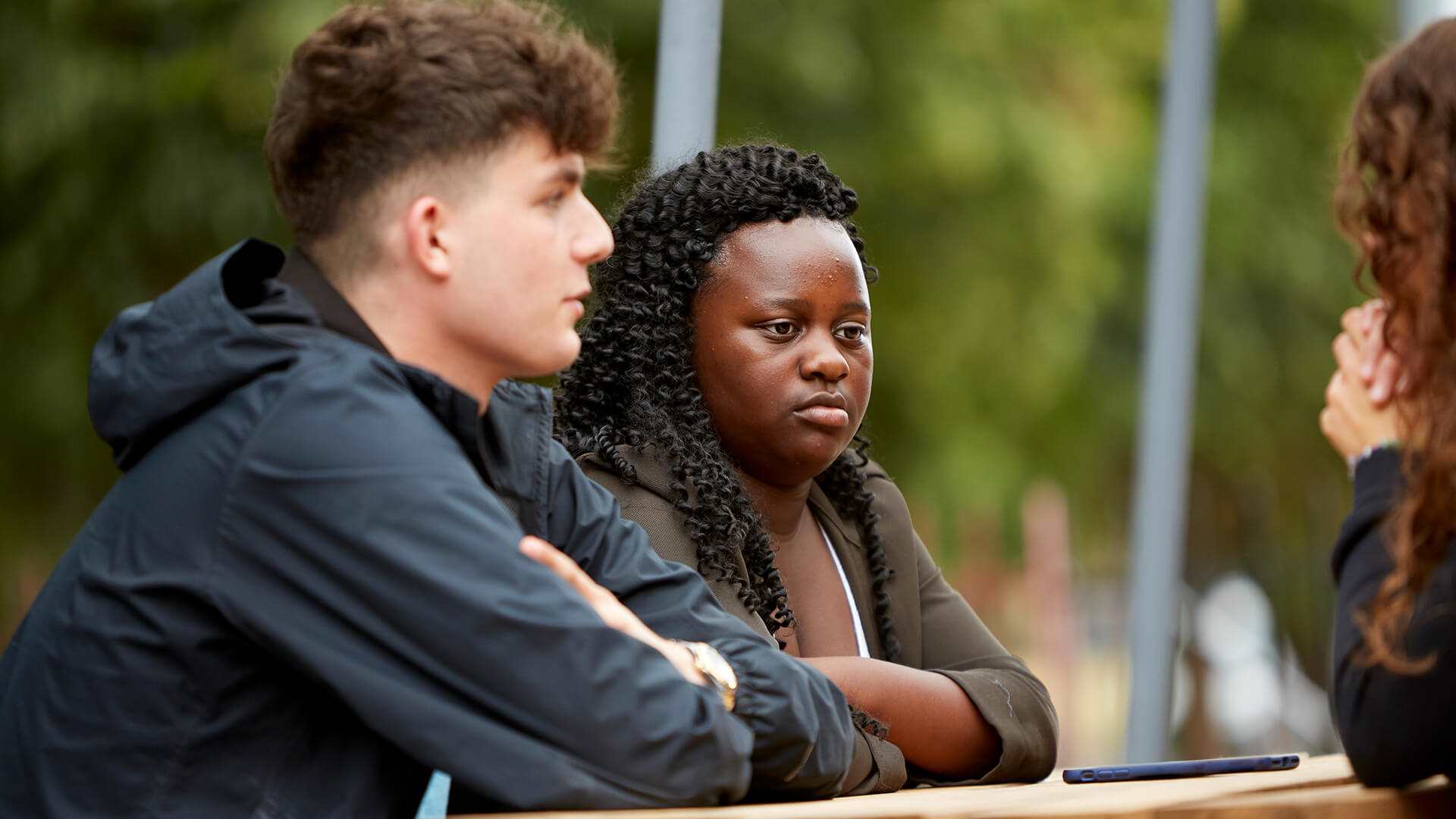 Two young people listening intently to their friend while sitting on a bench in a park.