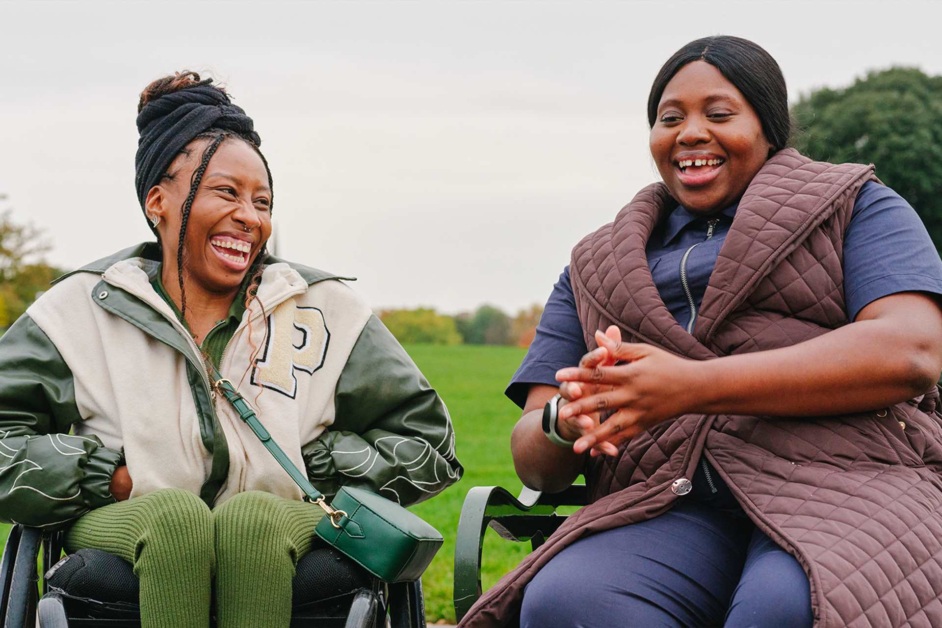 A young Black woman in a wheelchair and an older Black woman sitting on a bench in the park. They are laughing together.