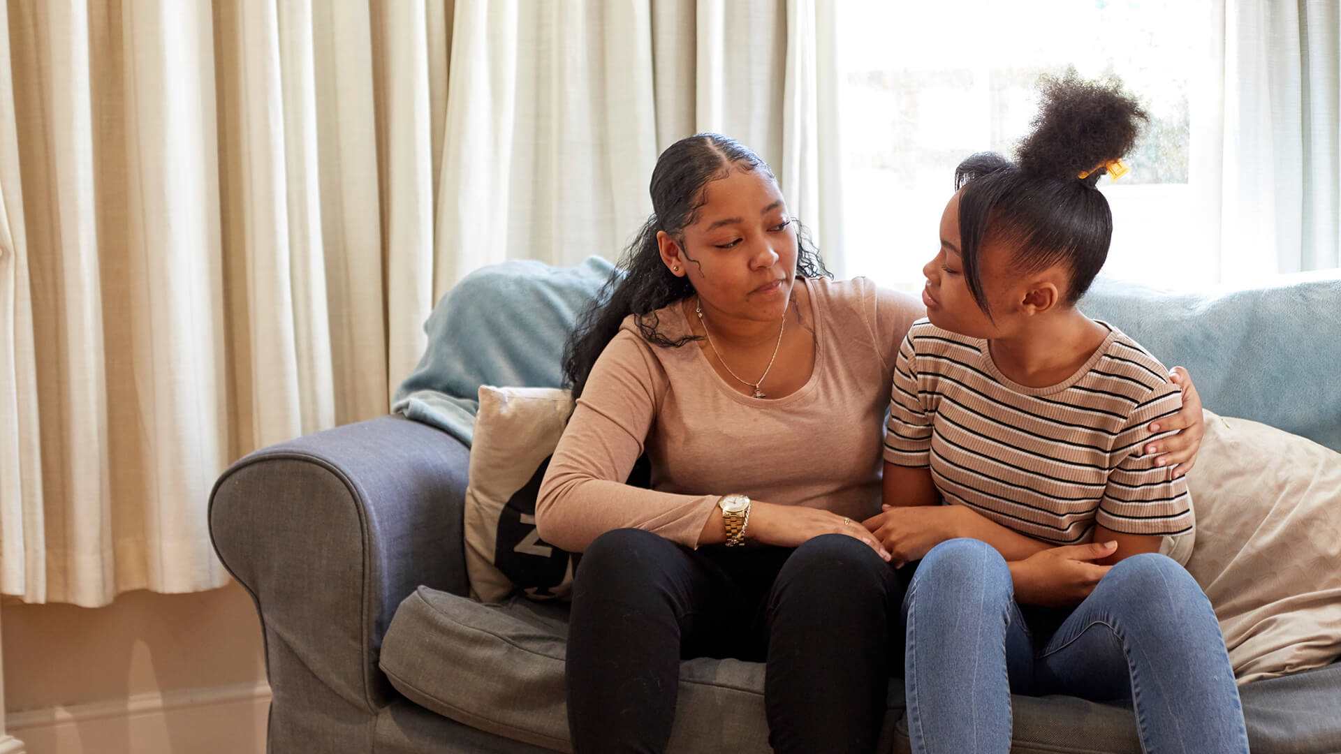 Two young people are sitting on a blue sofa facing each other. The person on the left has their arm around the person on the right.