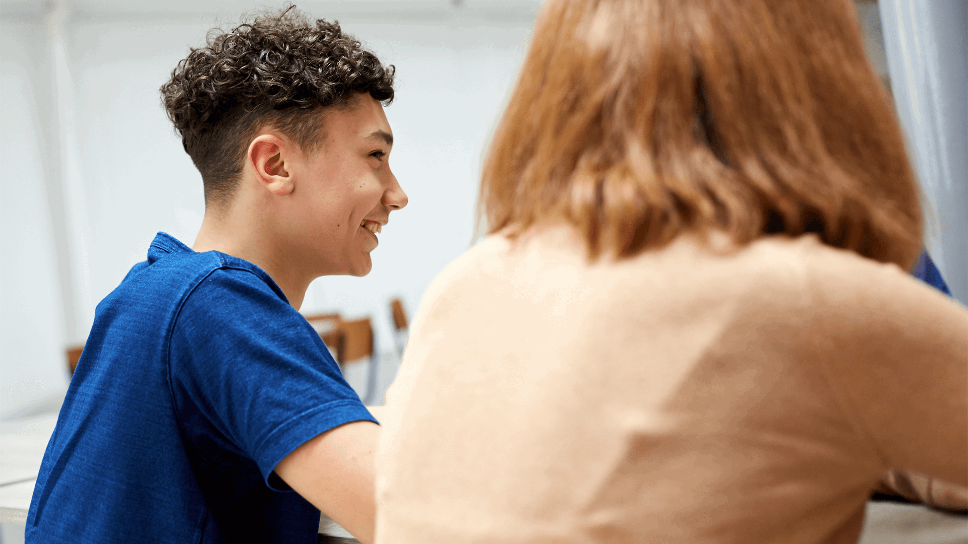 backshot-of-a-boy-with-short-curly-hair-and-wearing-blue-shirt-laughing-beside-a-girl-wearing-beige-shirt-with-face-unseen-while-inside-a-classroom