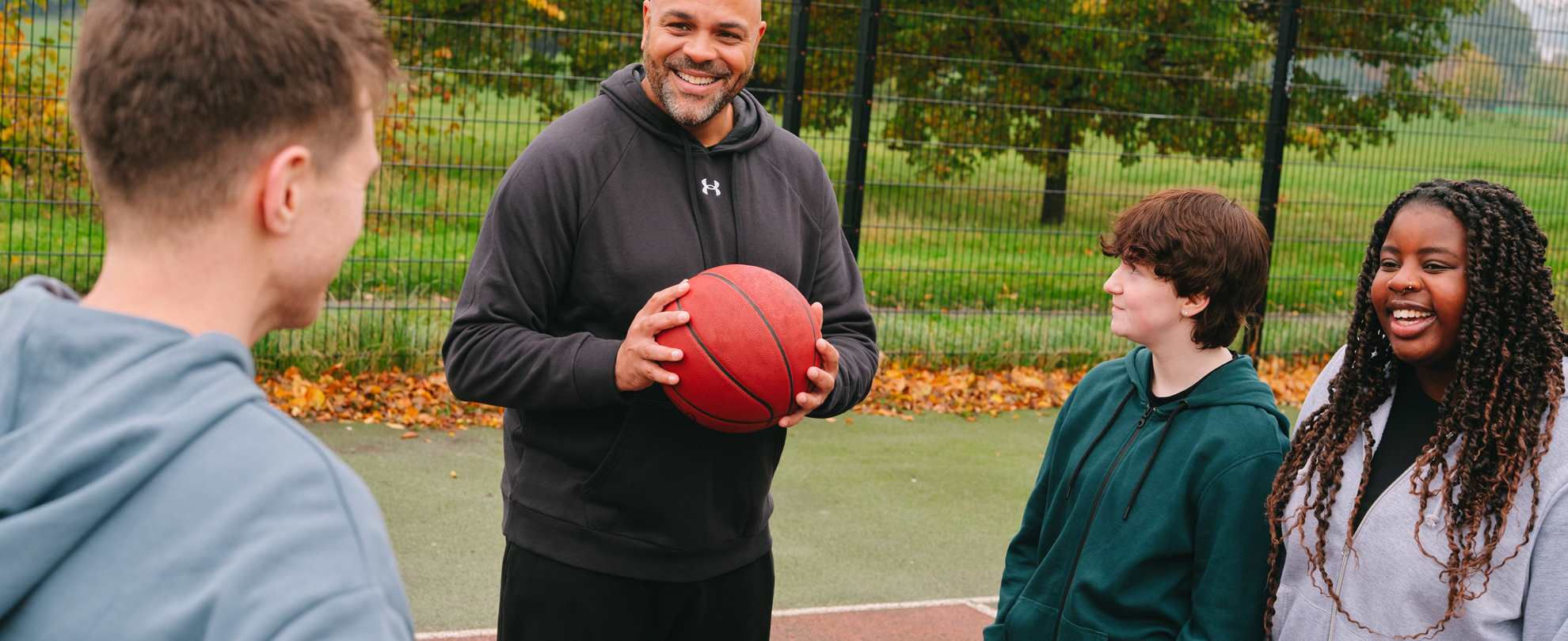 A group of young people playing basketball with an older Black man. The group of young people includes: one white young man, one white non-binary teenager and one Black young woman.