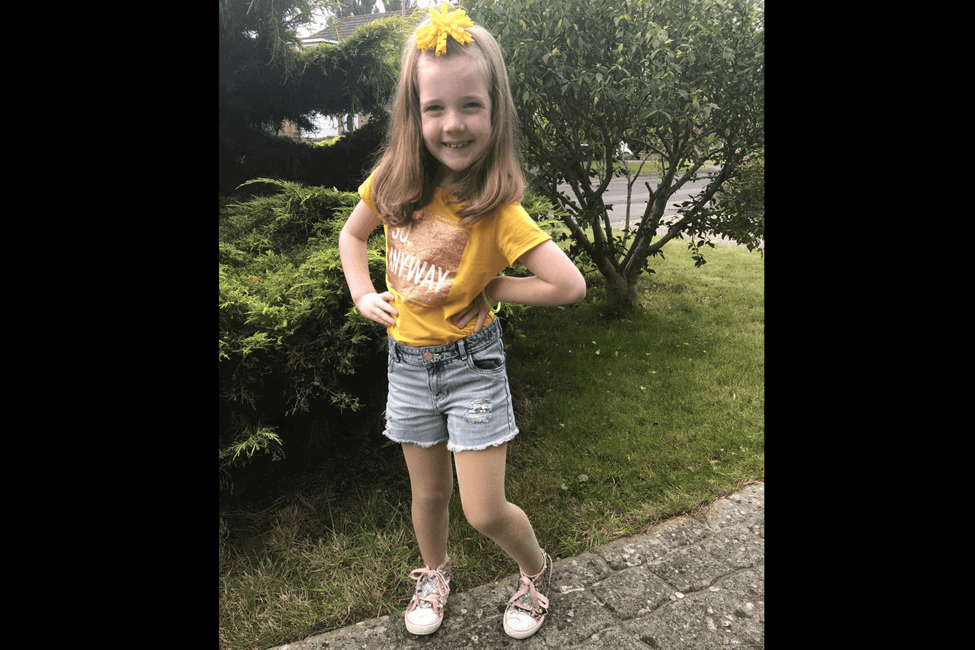 A young person stands with their hands on their hips wearing shorts, a yellow top and yellow bow in their hair.