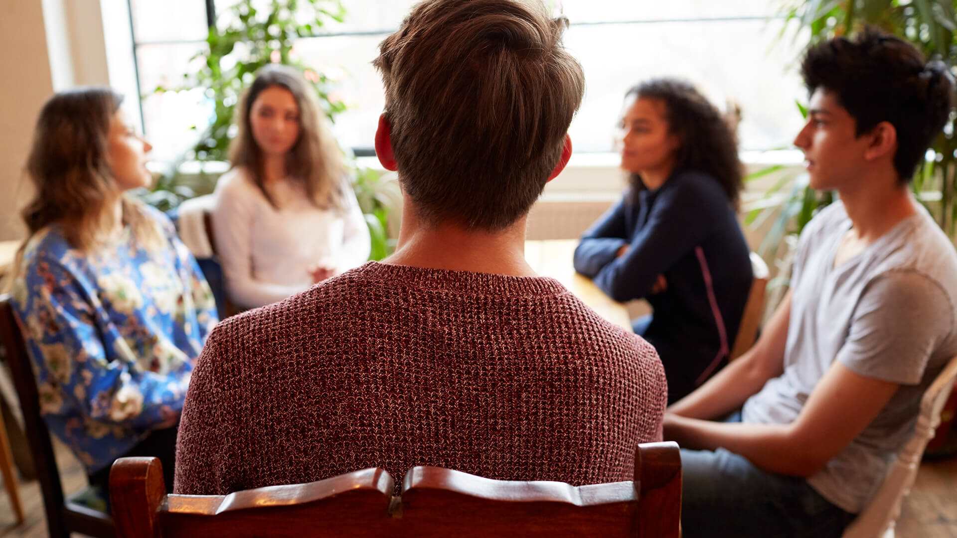 A group of young people sit around a table talking to each other. The person closest to the camera as the back to the camera and wears a dark red knitted jumper.