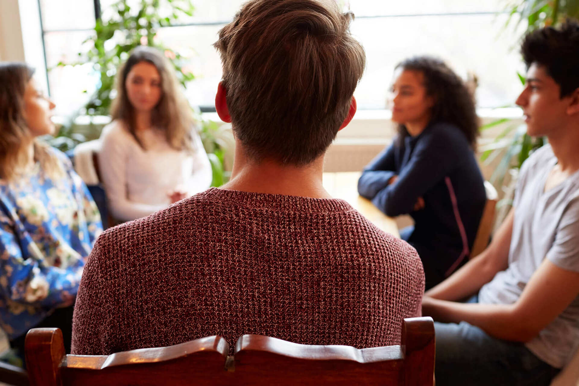 A group of young people sit around a table talking to each other. The person closest to the camera as the back to the camera and wears a dark red knitted jumper.