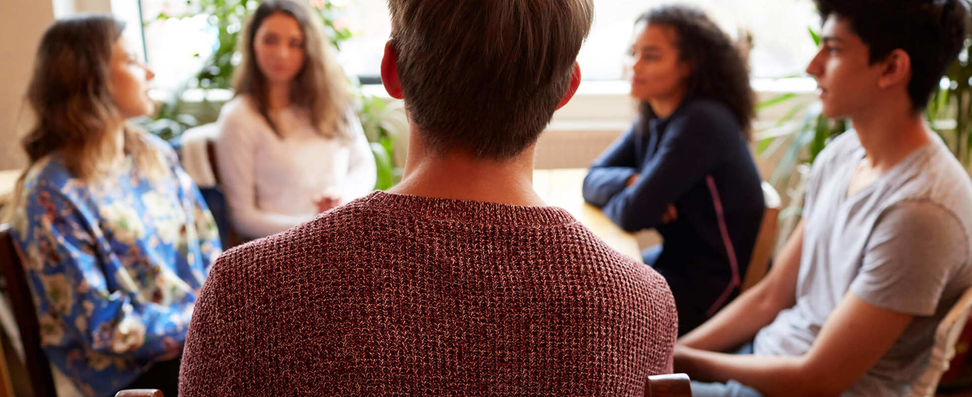 A group of young people sit around a table talking to each other. The person closest to the camera as the back to the camera and wears a dark red knitted jumper.