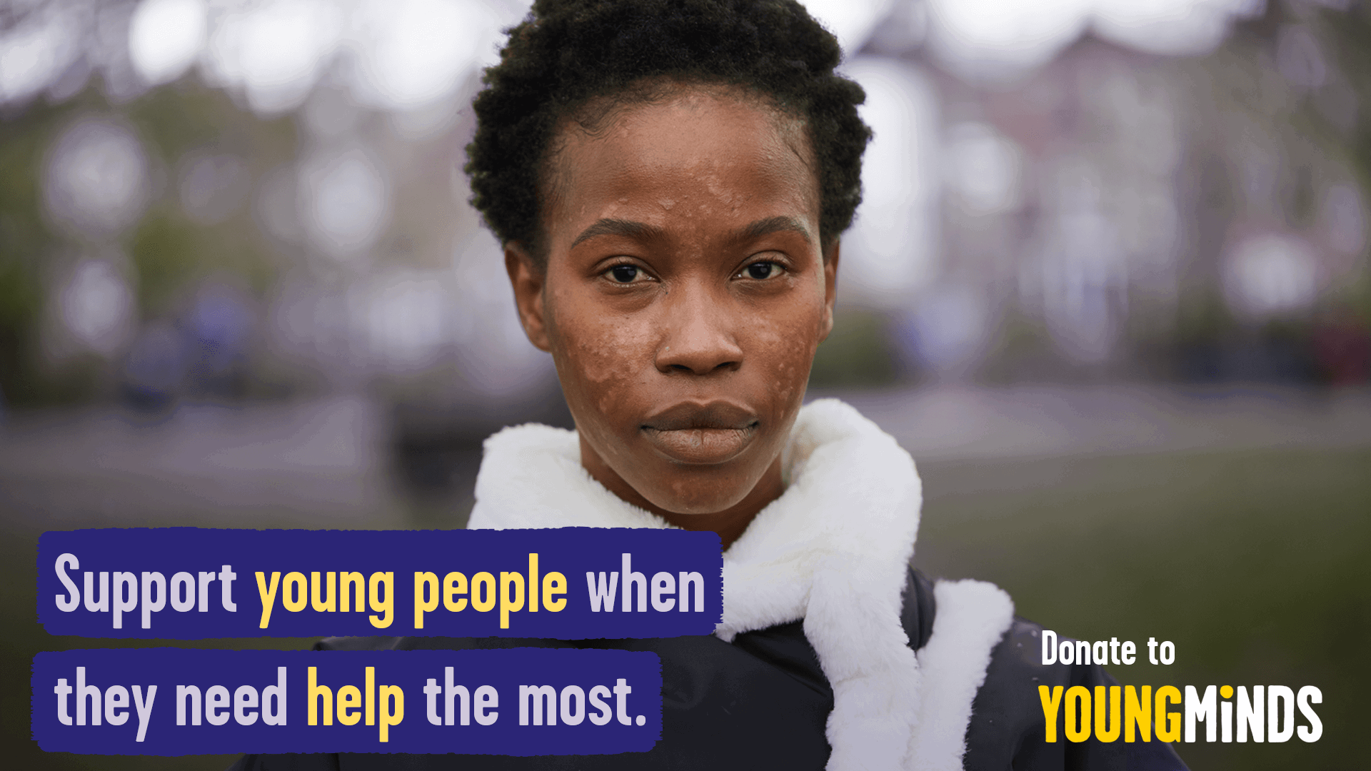 A young person is sitting on a bench, looking seriously straight at the camera. They wear a jacket with white fur. On the image are the words in purple and yellow 'support young people when they need help the most.' In the bottom right, it says 'Donate to YoungMinds'.