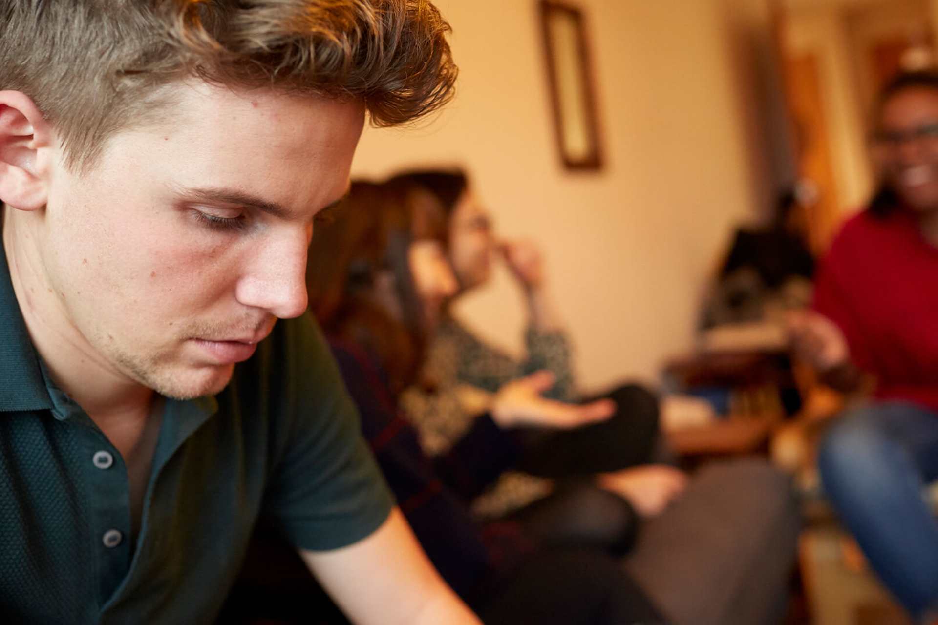 A young man looks worried and alone, while his three friends are talking and laughing in the background.