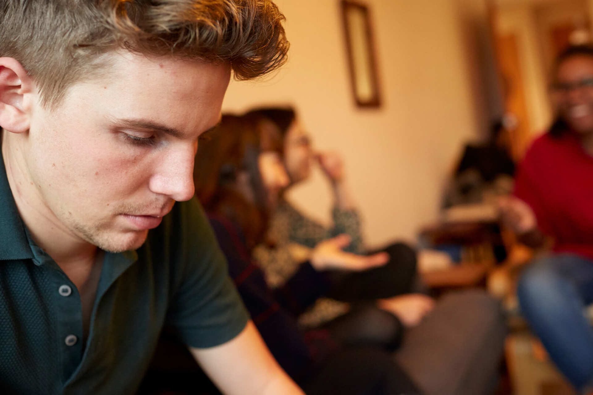 A young man looks worried and alone, while his three friends are talking and laughing in the background.