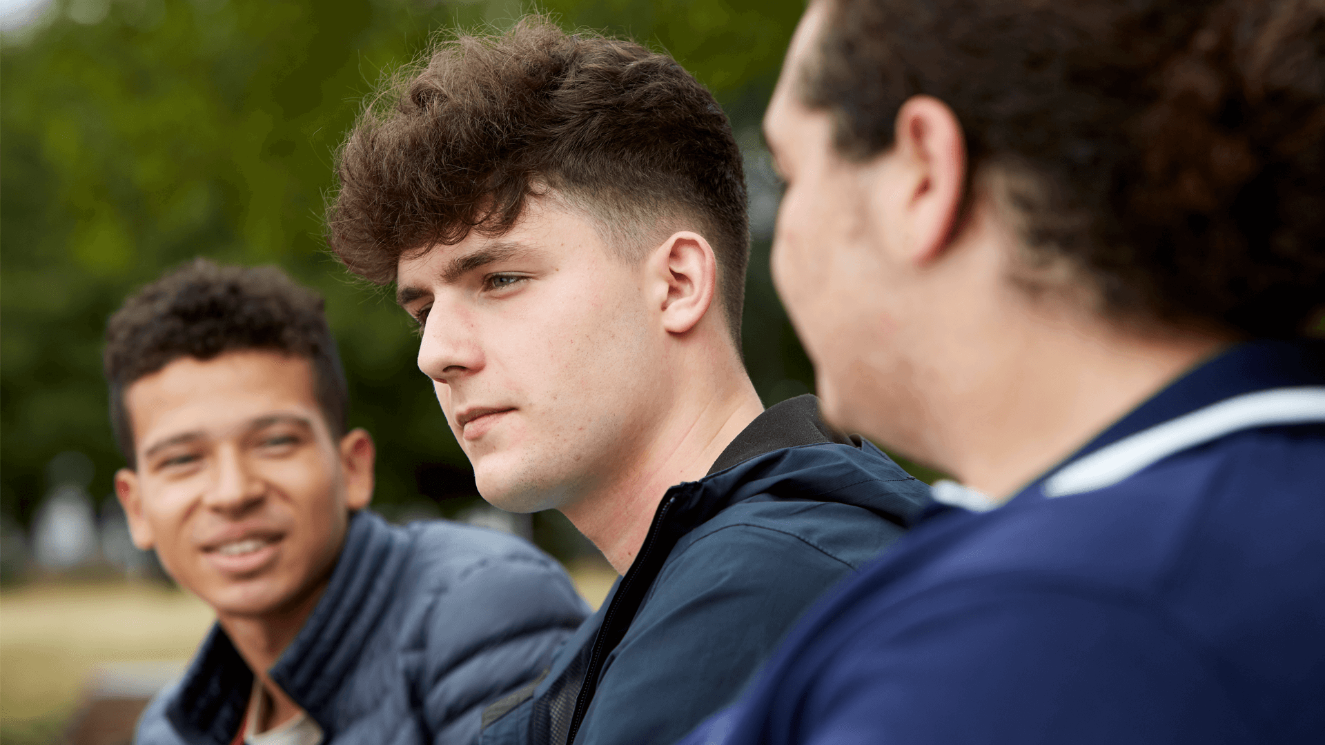 Three young people sitting together. One boy in the middle is looking away and seems to be deep in thought. The other two boys either side of him are looking at each other and talking.