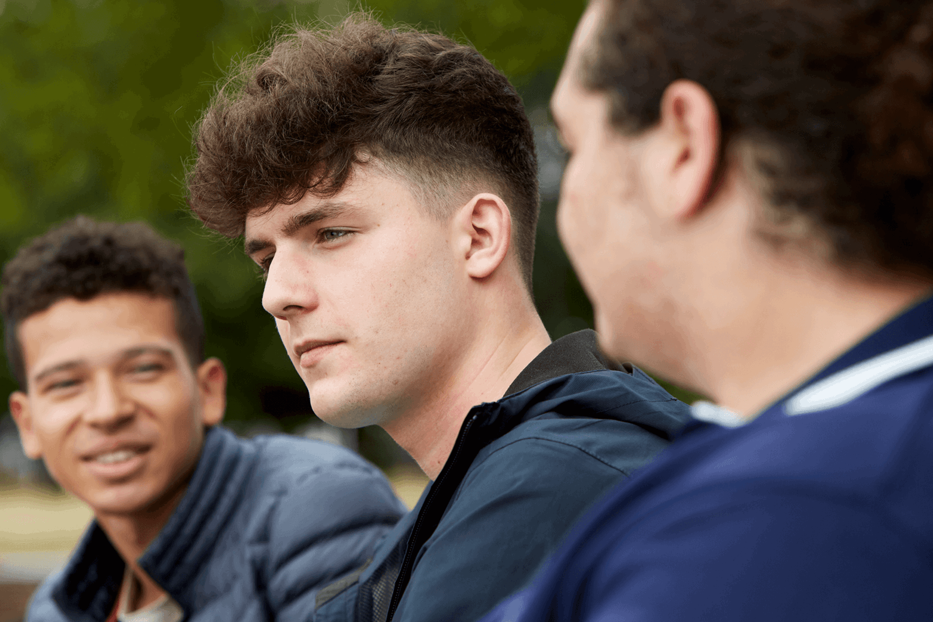 Three young people sitting together. One boy in the middle is looking away and seems to be deep in thought. The other two boys either side of him are looking at each other and talking.