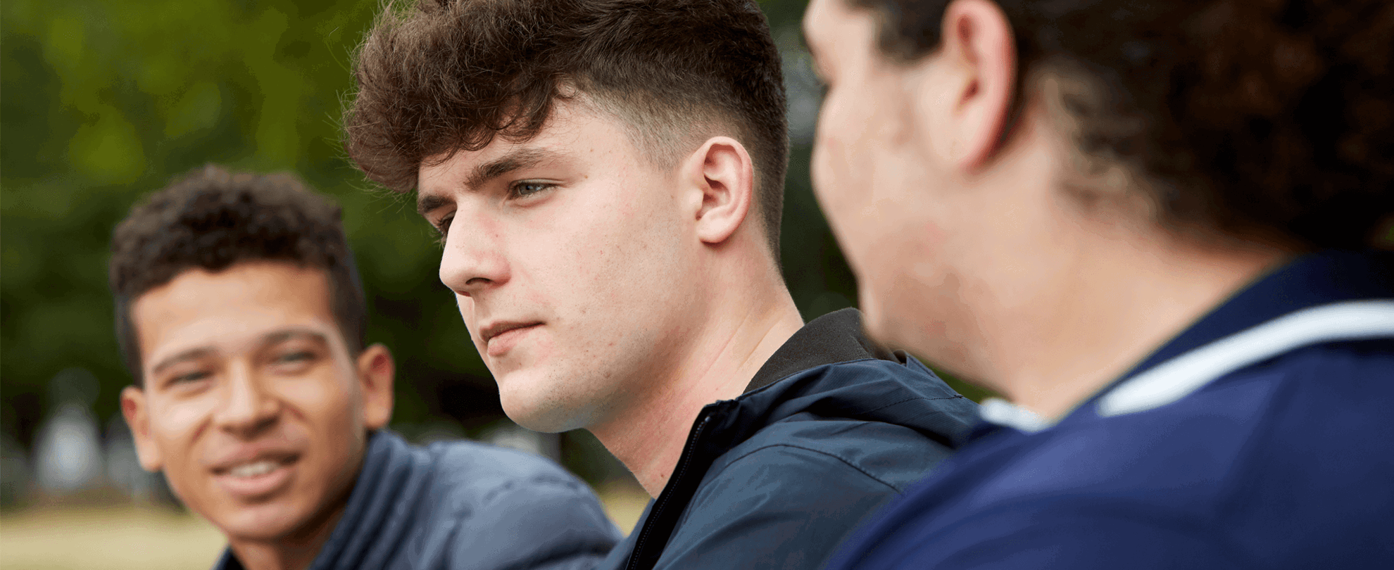 Three young people sitting together. One boy in the middle is looking away and seems to be deep in thought. The other two boys either side of him are looking at each other and talking.