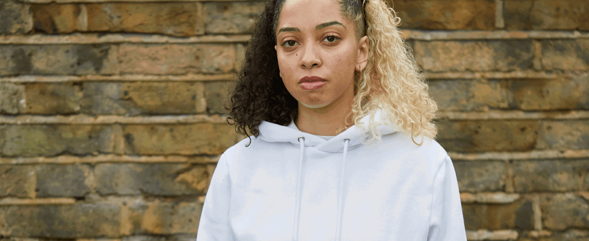 medium shot of a girl with curly hair wearing white hoodie looking in front of the camera with brick wall on the background