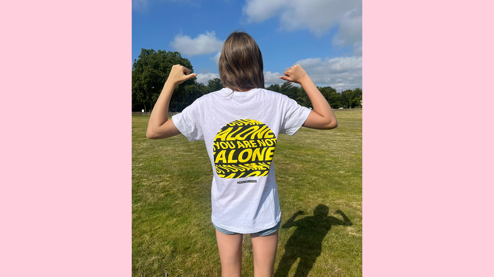 A person standing in a field with their back to the camera. They are wearing a YoungMinds t-shirt and pointing their thumbs to the logo over their shoulders.