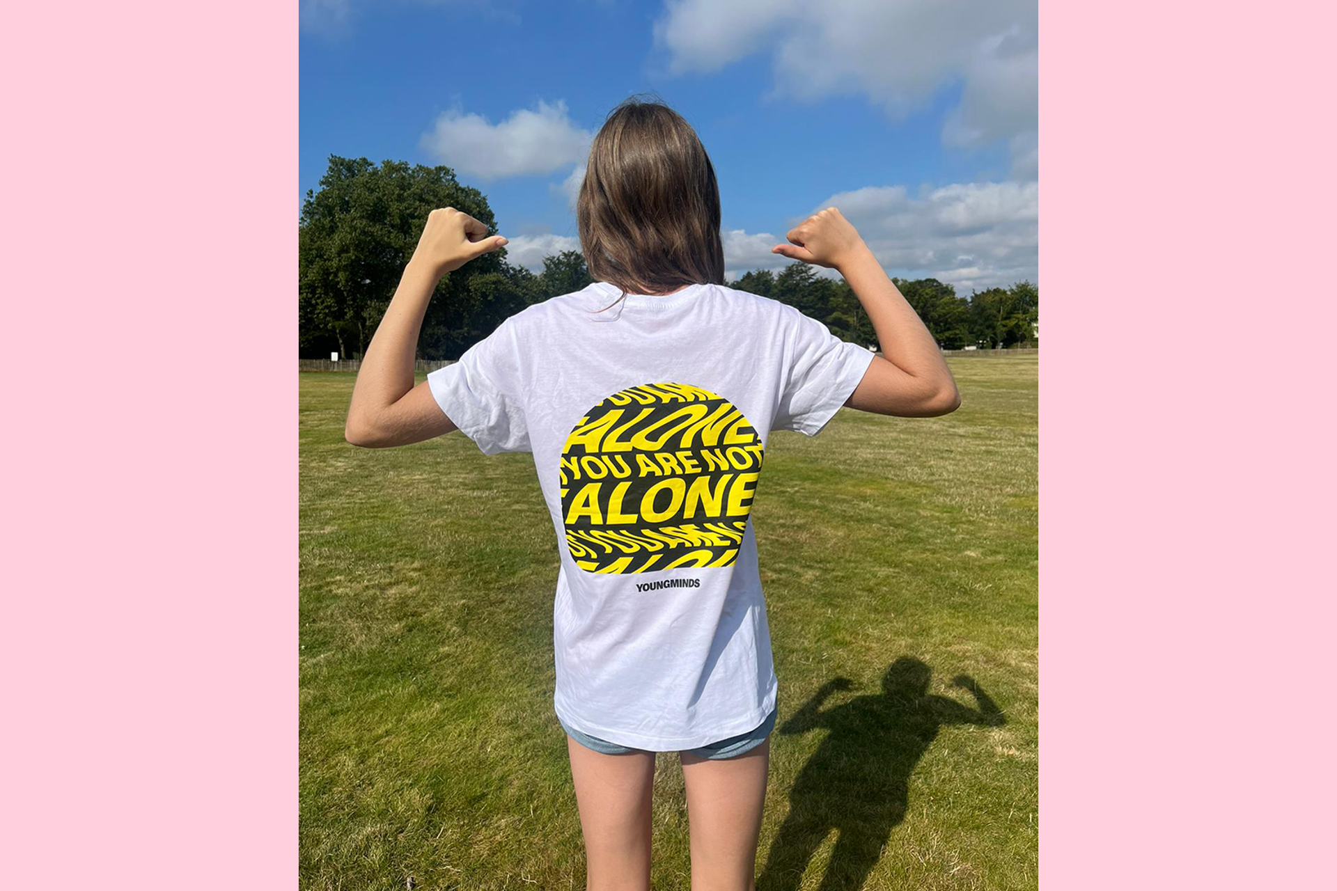 A person standing in a field with their back to the camera. They are wearing a YoungMinds t-shirt and pointing their thumbs to the logo over their shoulders.