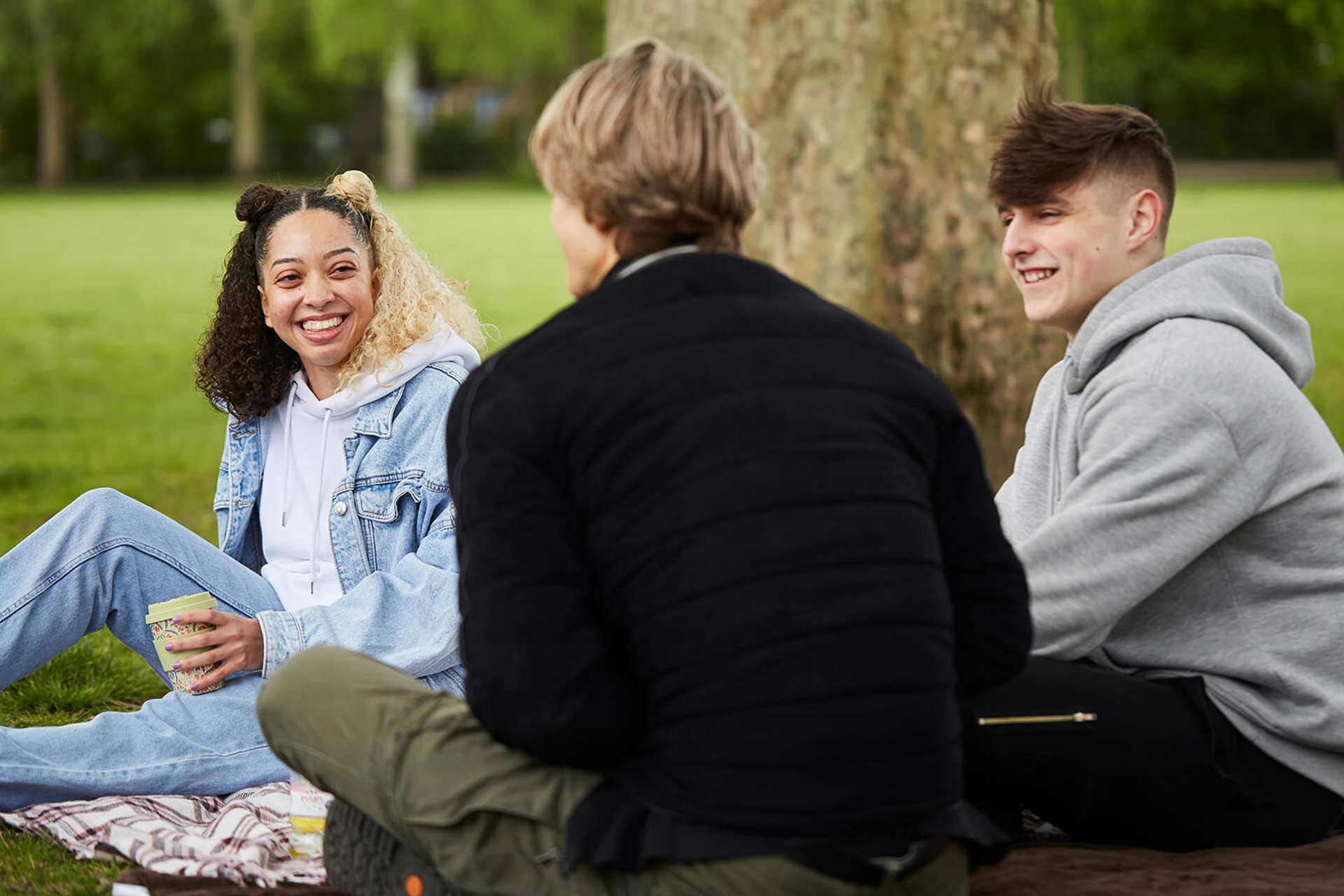 A group of three young people laugh and chat while sitting on the ground beside a tree in the park.