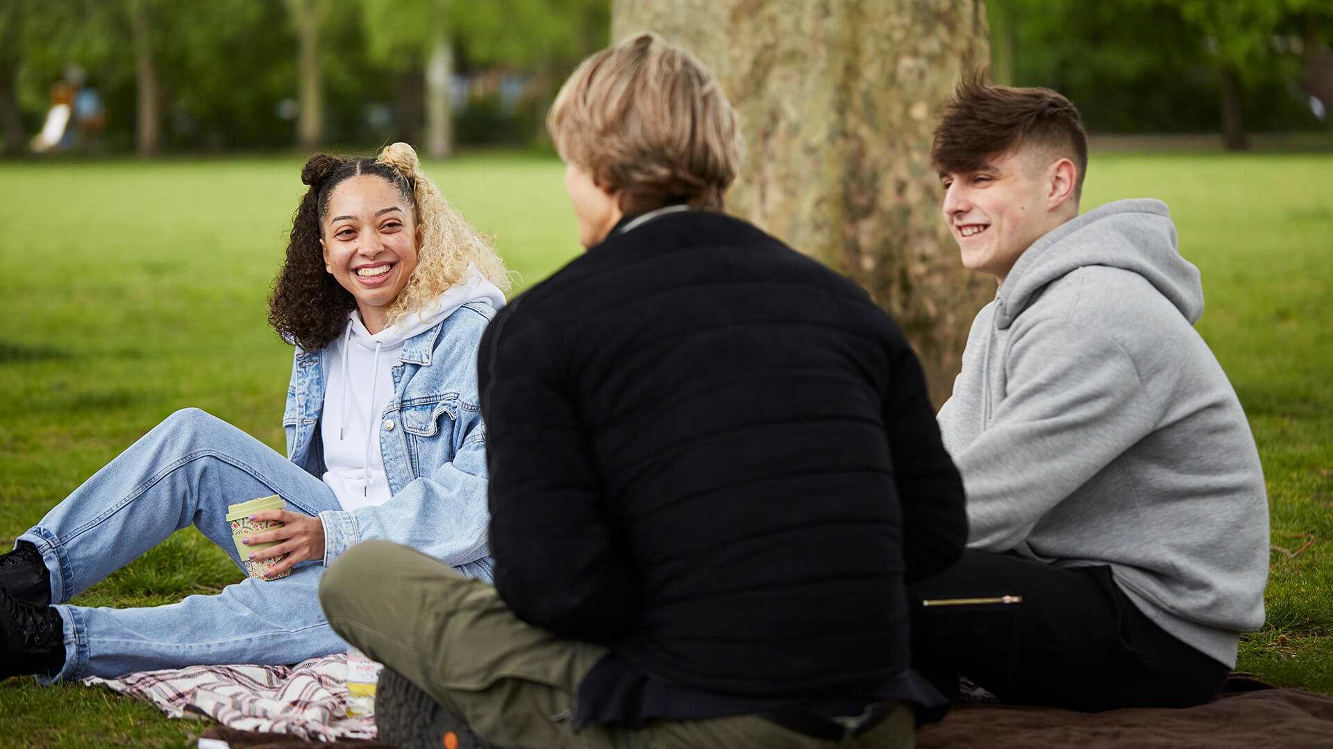 A group of three young people laugh and chat while sitting on the ground beside a tree in the park.
