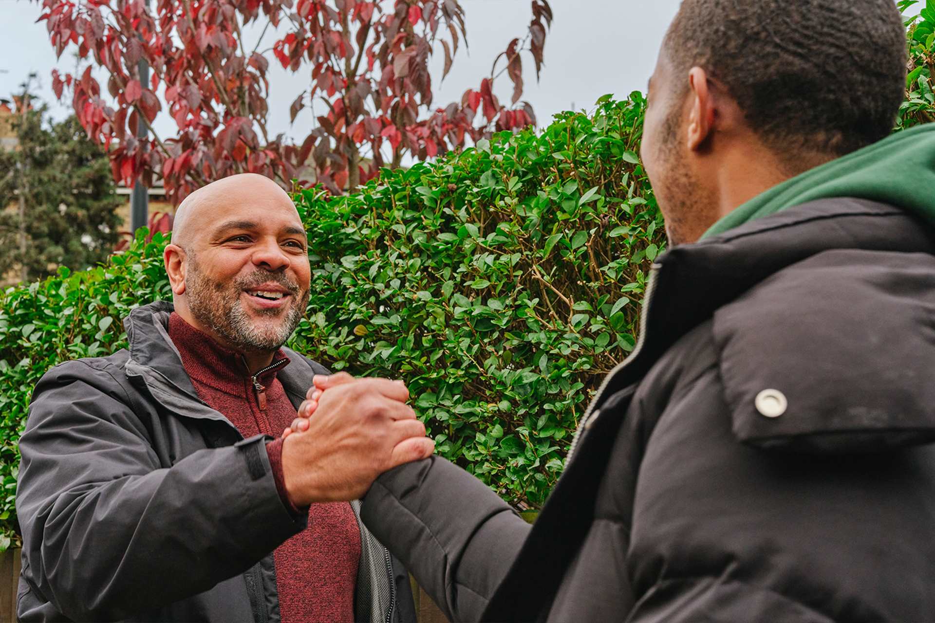 A young Black man grasping hands with an older Black man on the street. They are both smiling.