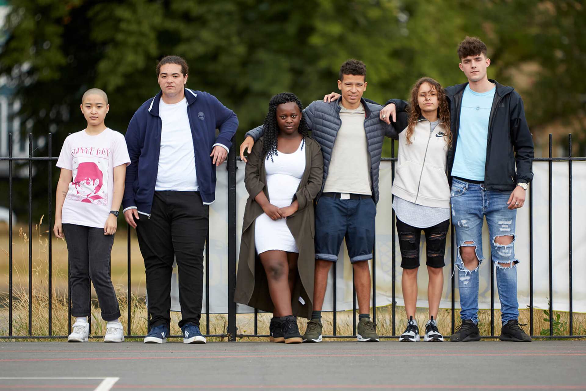 A mixed group of six young people standing in front of a black metal fence in a park playground.