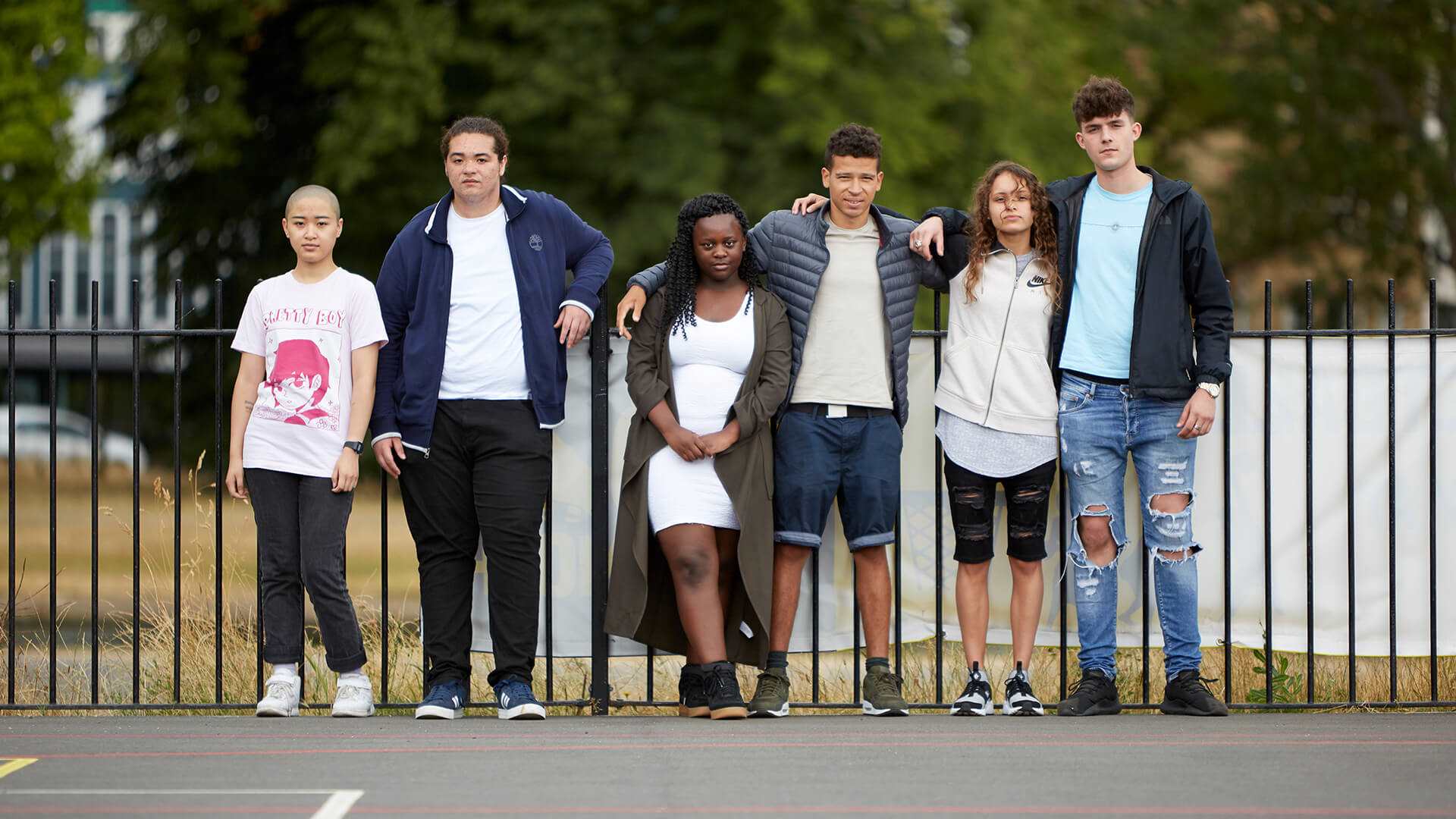 A mixed group of six young people standing in front of a black metal fence in a park playground.
