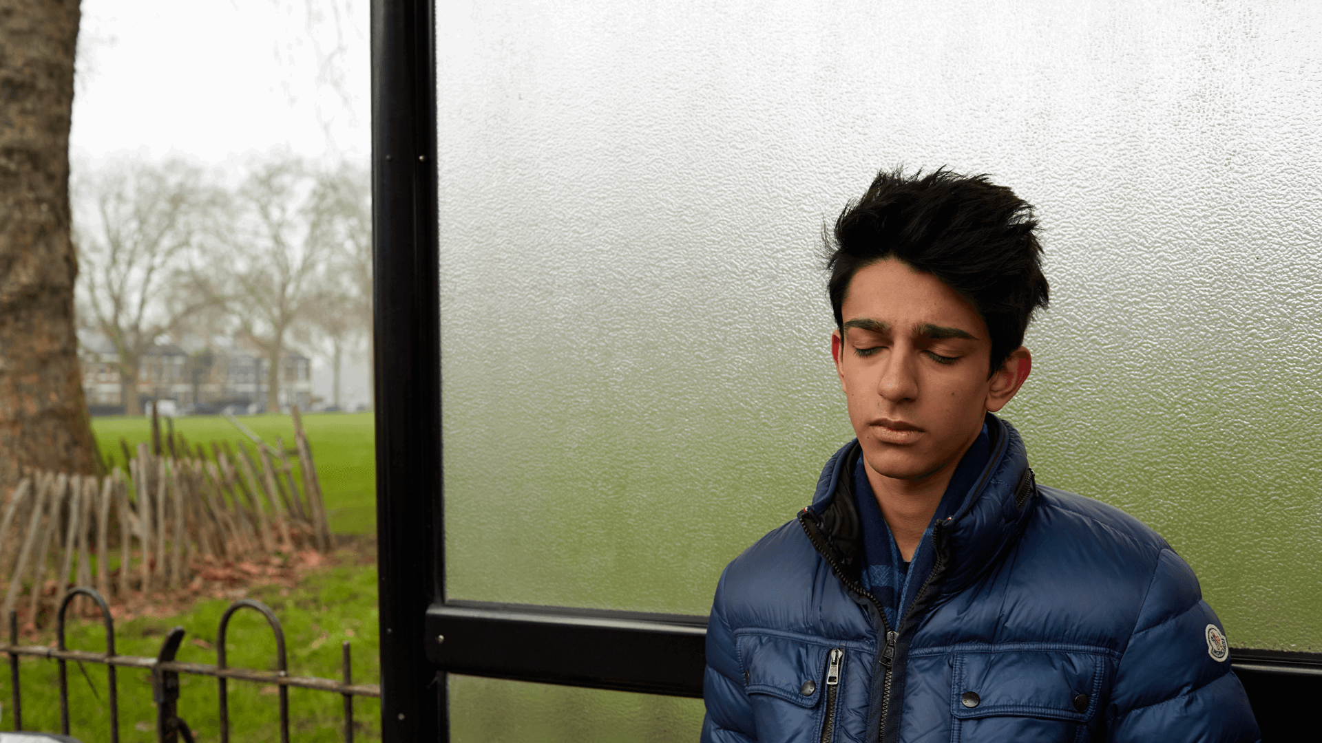 A boy wearing a blue jacket, standing alone a bus shelter and thinking with his eyes closed.