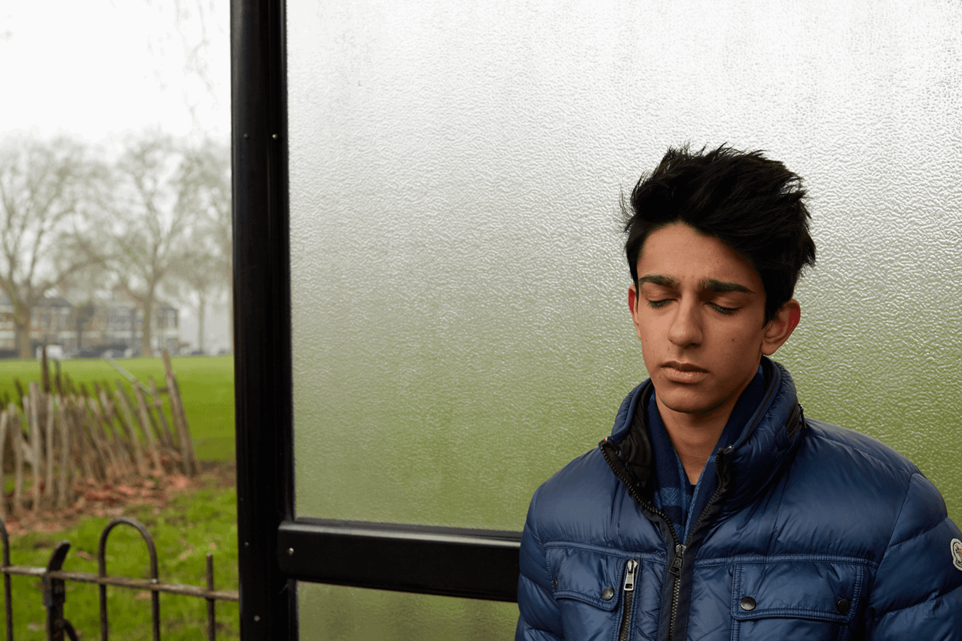 A boy wearing a blue jacket, standing alone a bus shelter and thinking with his eyes closed.