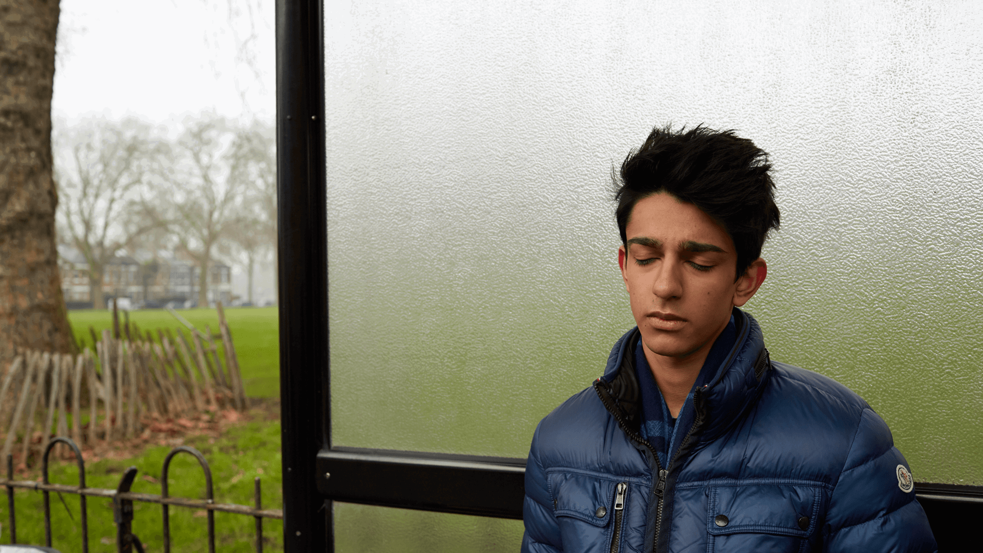 A boy wearing a blue jacket, standing alone a bus shelter and thinking with his eyes closed.