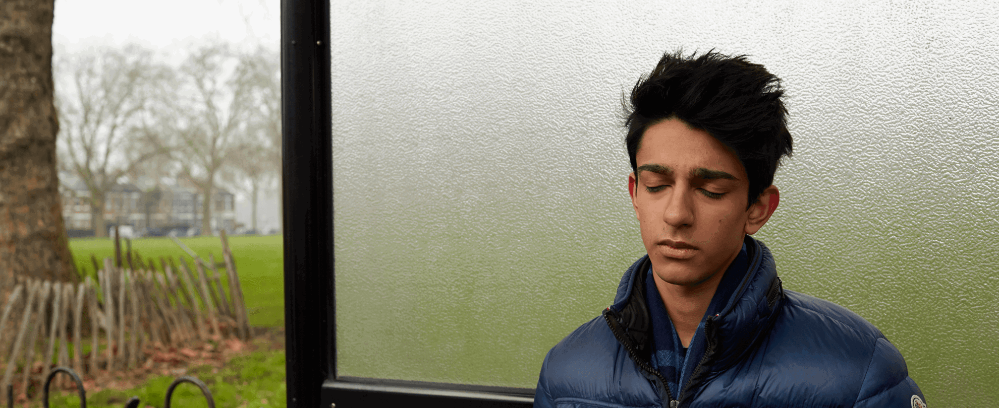 A boy wearing a blue jacket, standing alone a bus shelter and thinking with his eyes closed.