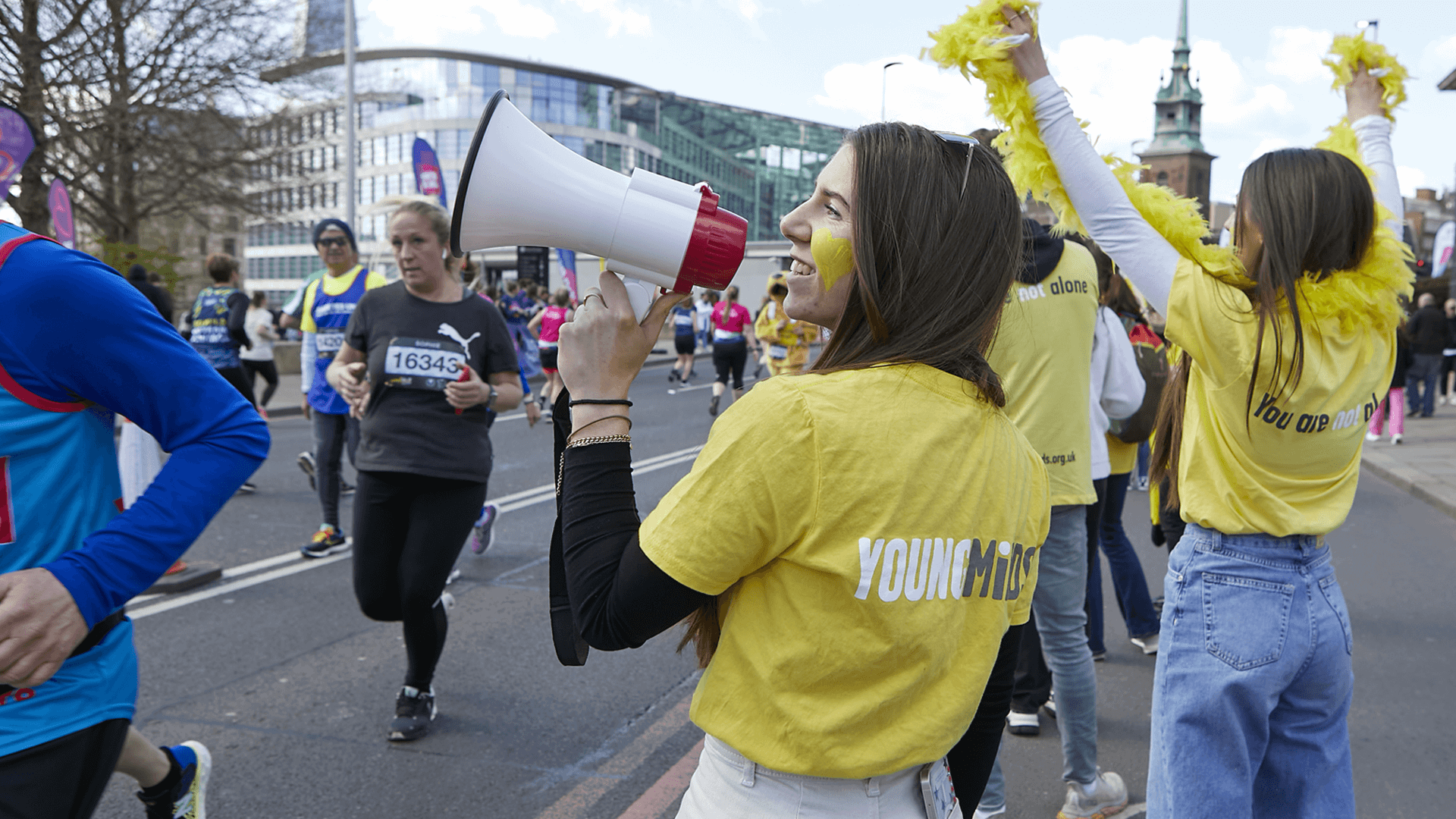 Volunteers in YoungMinds tops cheer passing runners at a half marathon, with a megaphone and feather boa.