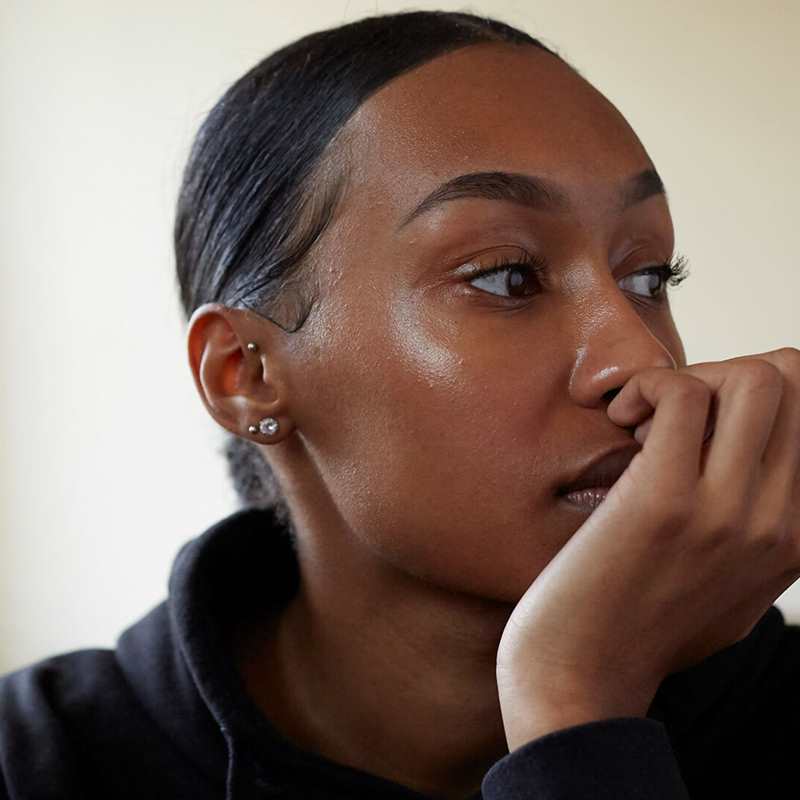 A young person sits in a room wearing a black hoodie and their hair tied back. They are looking to the right with their hand curled over their mouth, lost in thought.