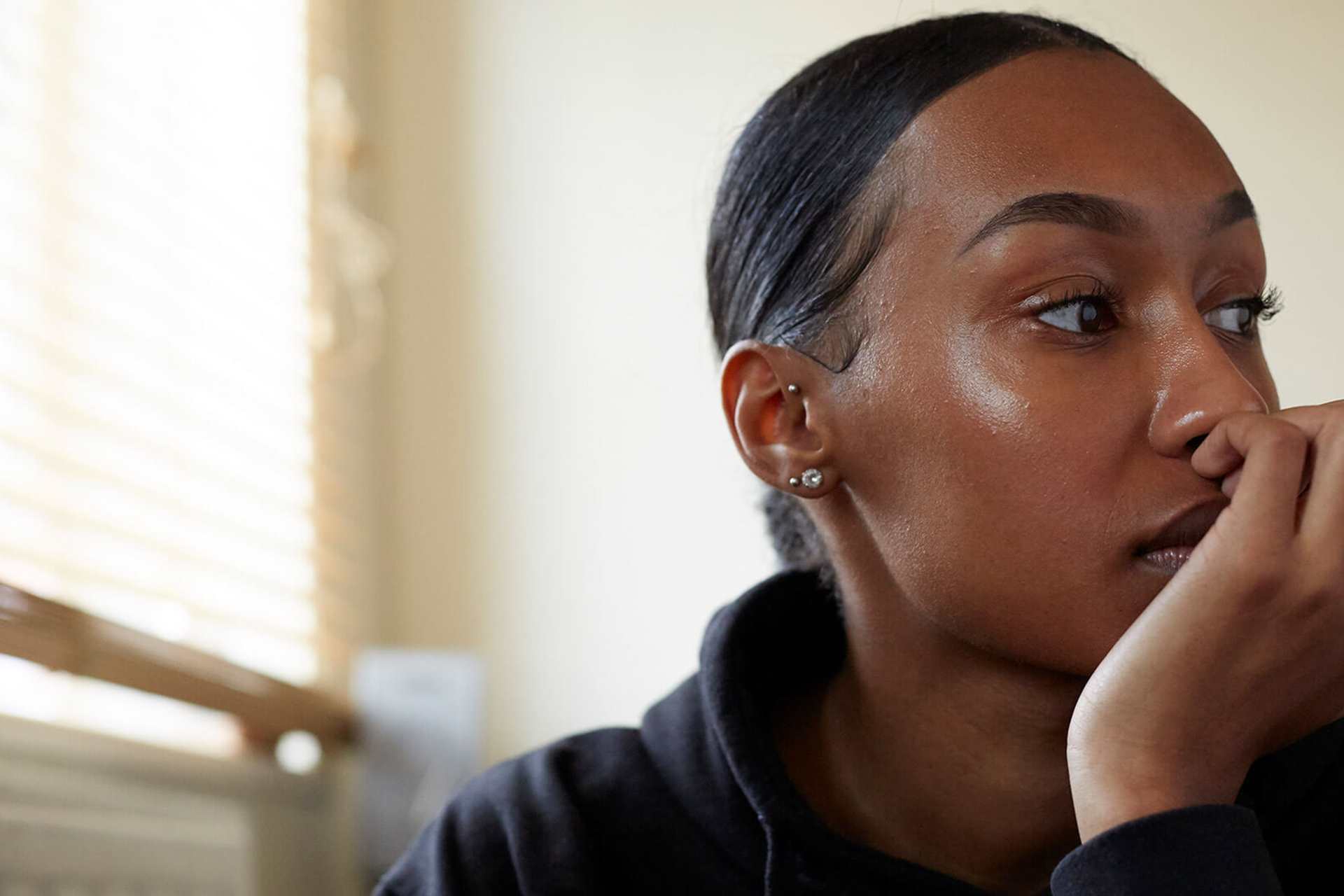 A young person sits in a room wearing a black hoodie and their hair tied back. They are looking to the right with their hand curled over their mouth, lost in thought.