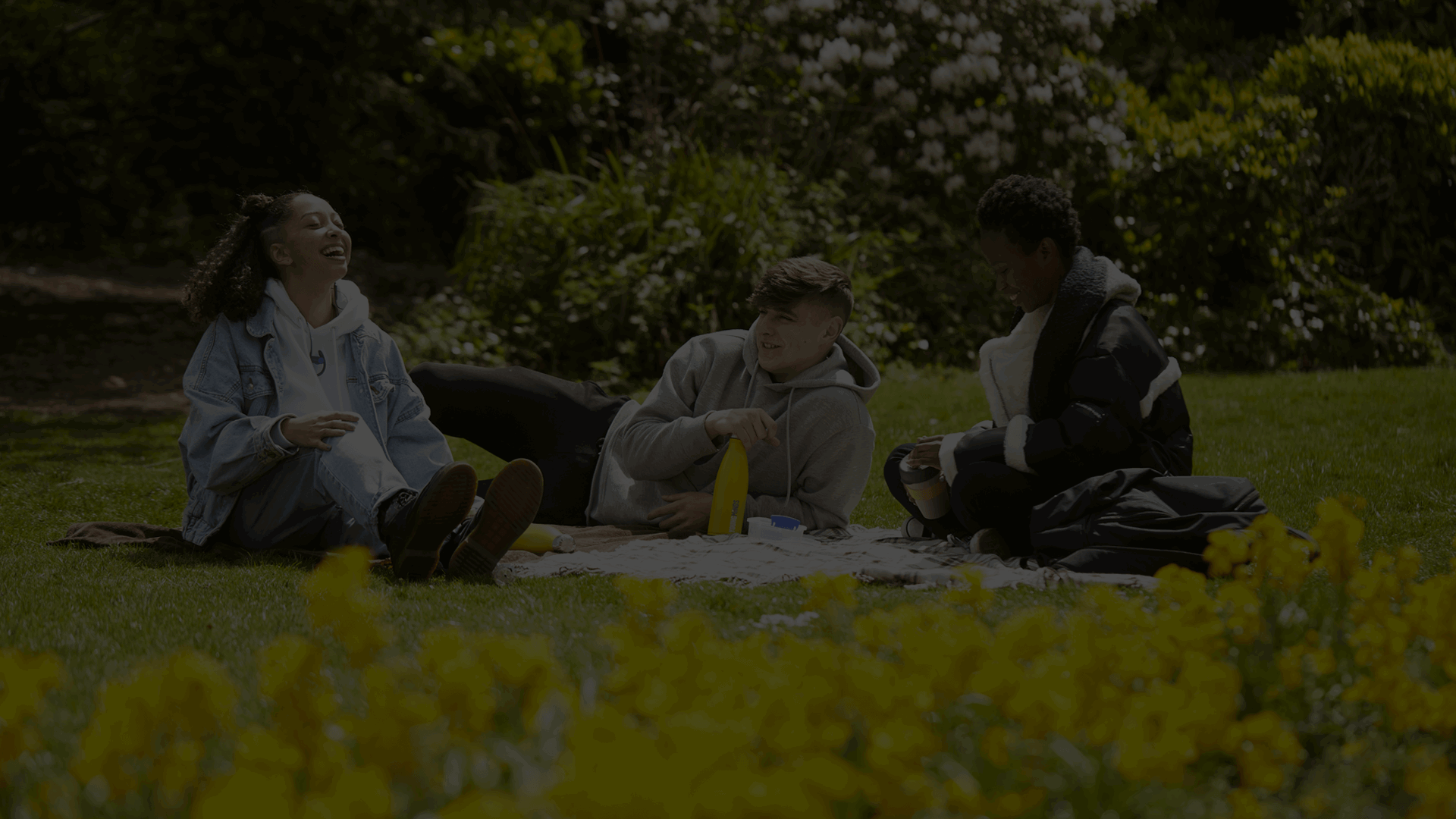 Three young people sit outside on the grass with a picnic blanket. There are bushes and flowers around them with some yellow flowers in front of the camera. The person on the left is looking up and laughing, the person in the middle holds a YoungMinds yellow water bottle while looking at the person laughing. The person on the right sits looking down at their hands while smiling.