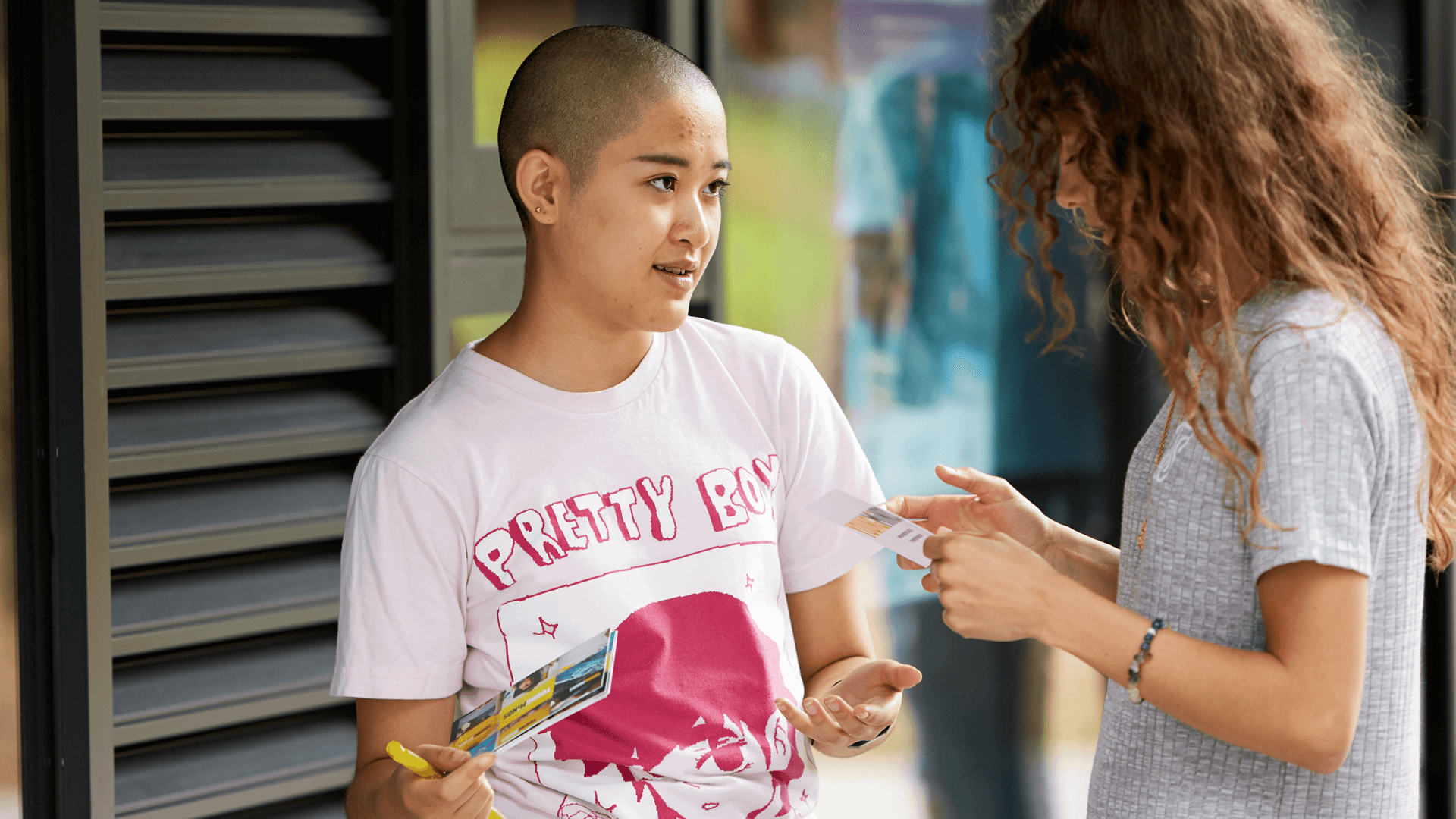 medium-shot-of-a-young-woman-with-shaved-head-holding-some-flyers-talking-to-another-girl-with-curly-hair-and-face-unseen-inside-campus