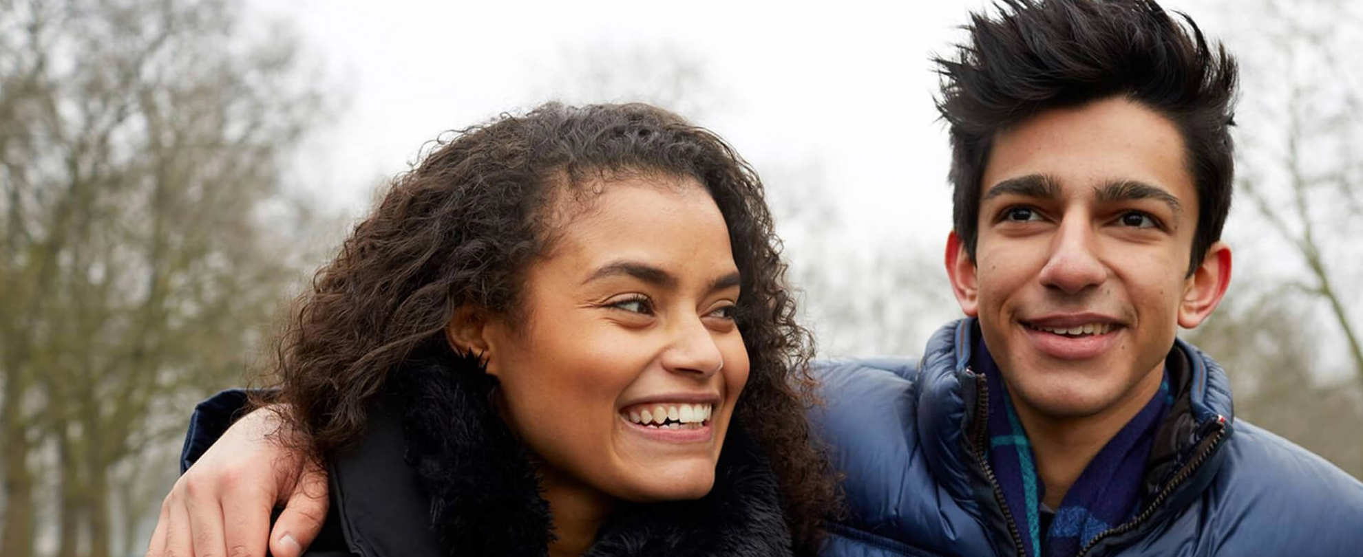 Two young people standing together and smiling, one has their arm around the shoulder of the other.