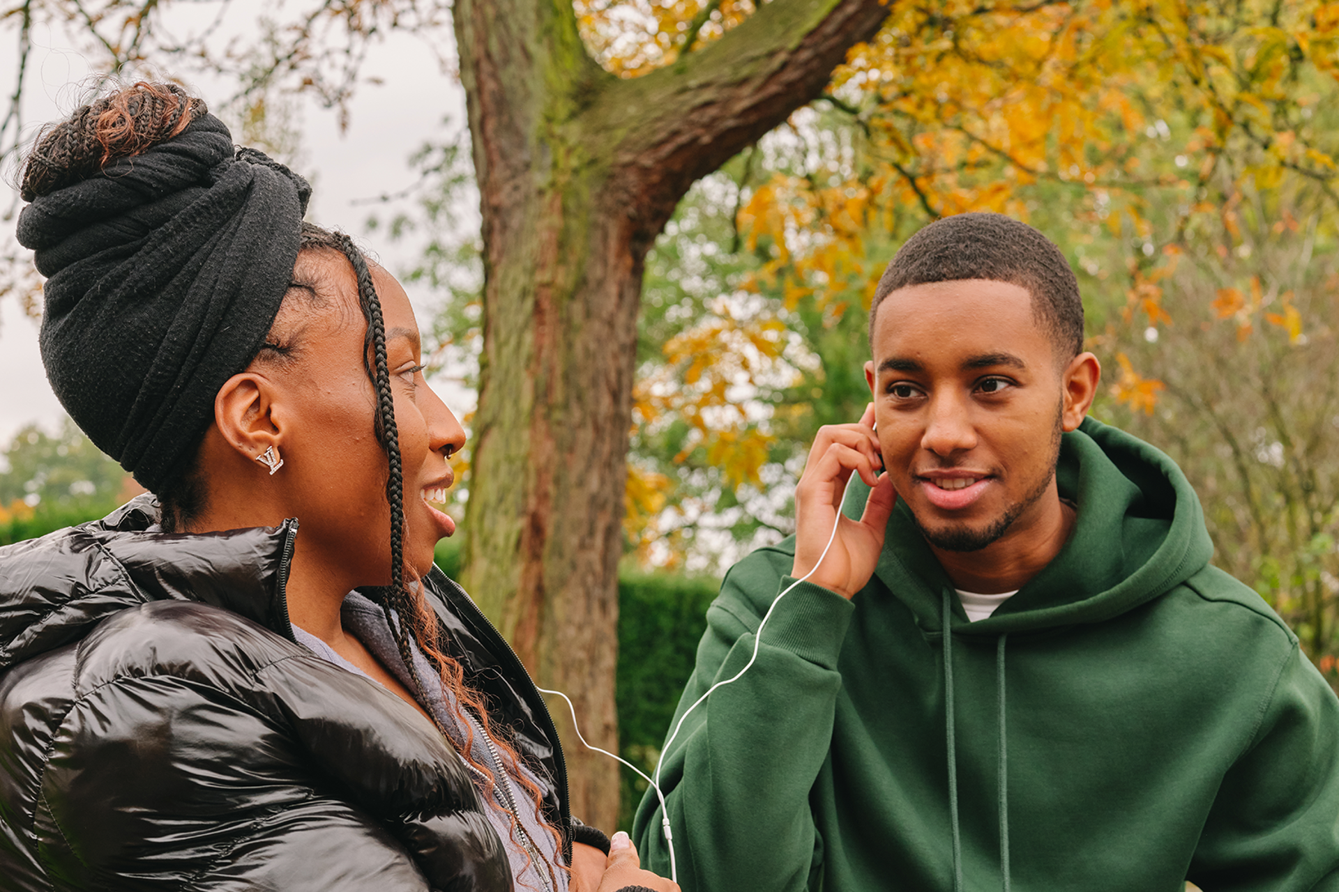 A Black young woman listening to music through headphones with a Black young man in the park.