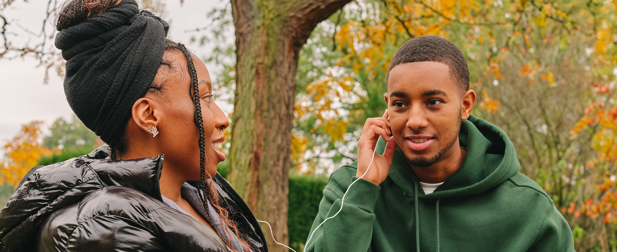 A Black young woman listening to music through headphones with a Black young man in the park.