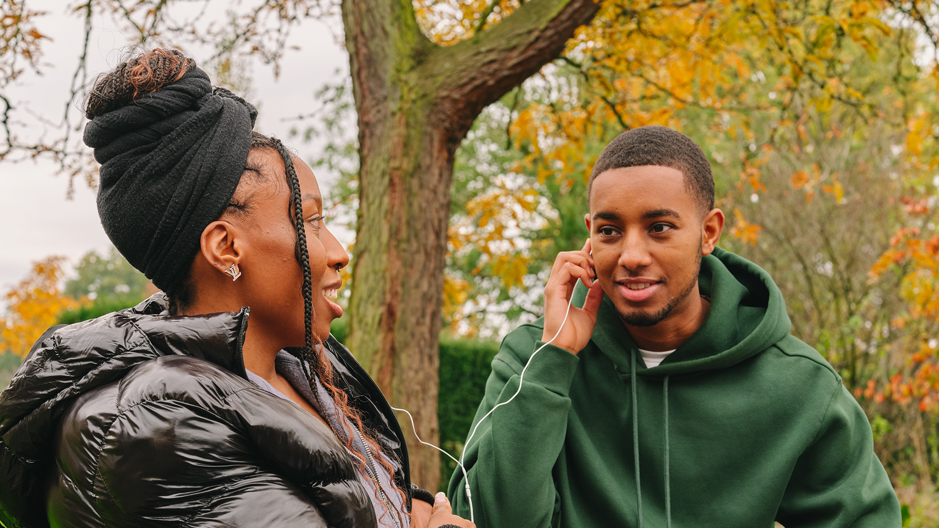 A Black young woman listening to music through headphones with a Black young man in the park.