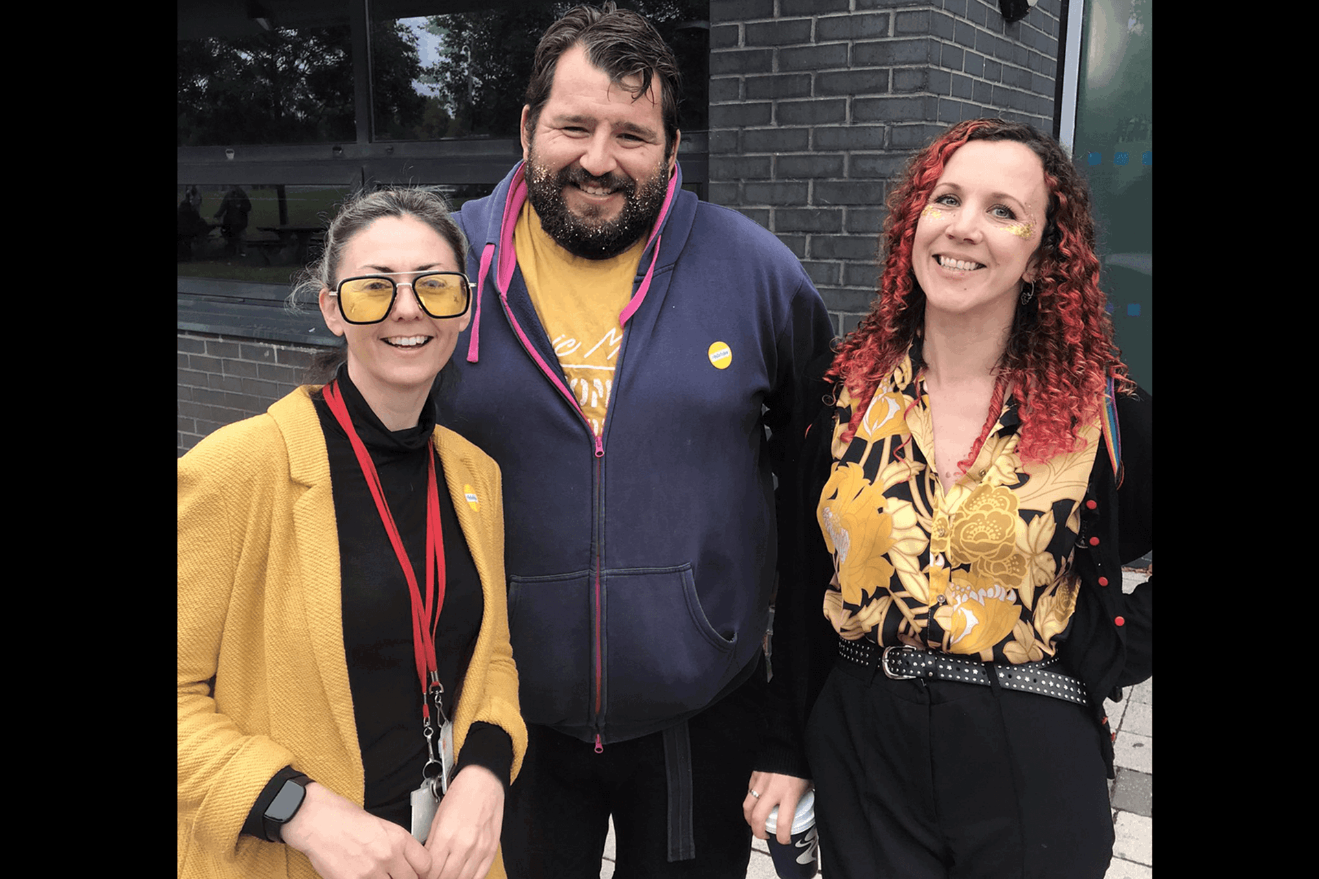 Three people stand together smiling. The person on the left wears a yellow coat and sunglasses. The person in the middle has a yellow top and yellow glitter beard and the person on the right is wearing a yellow flower shirt.