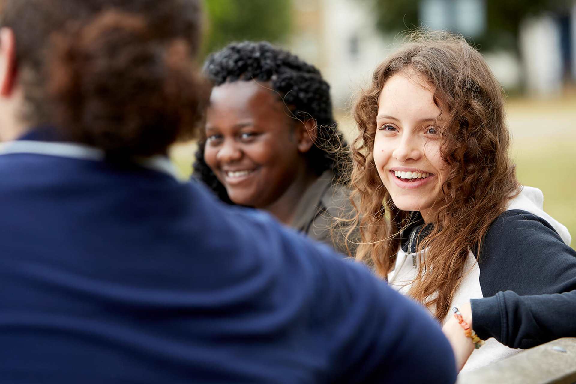 Three young people sit on a bench talking and laughing. The person on the left has their back to the camera and wears a dark blue jacket. The person in the middle has long wavy hair and wears a black and white jacket. The person on the right has curly hair and wears a green jacket.