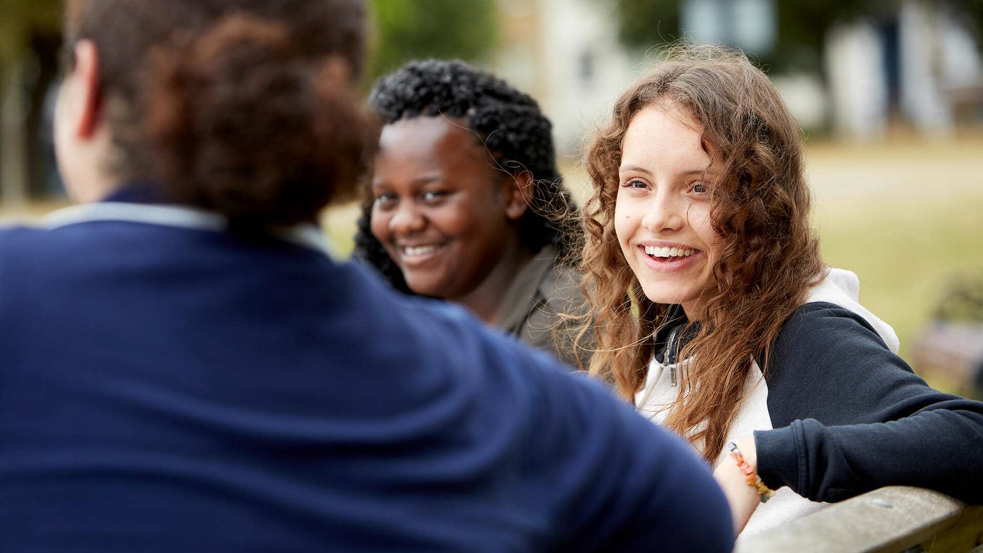 Three young people sit on a bench talking and laughing. The person on the left has their back to the camera and wears a dark blue jacket. The person in the middle has long wavy hair and wears a black and white jacket. The person on the right has curly hair and wears a green jacket.