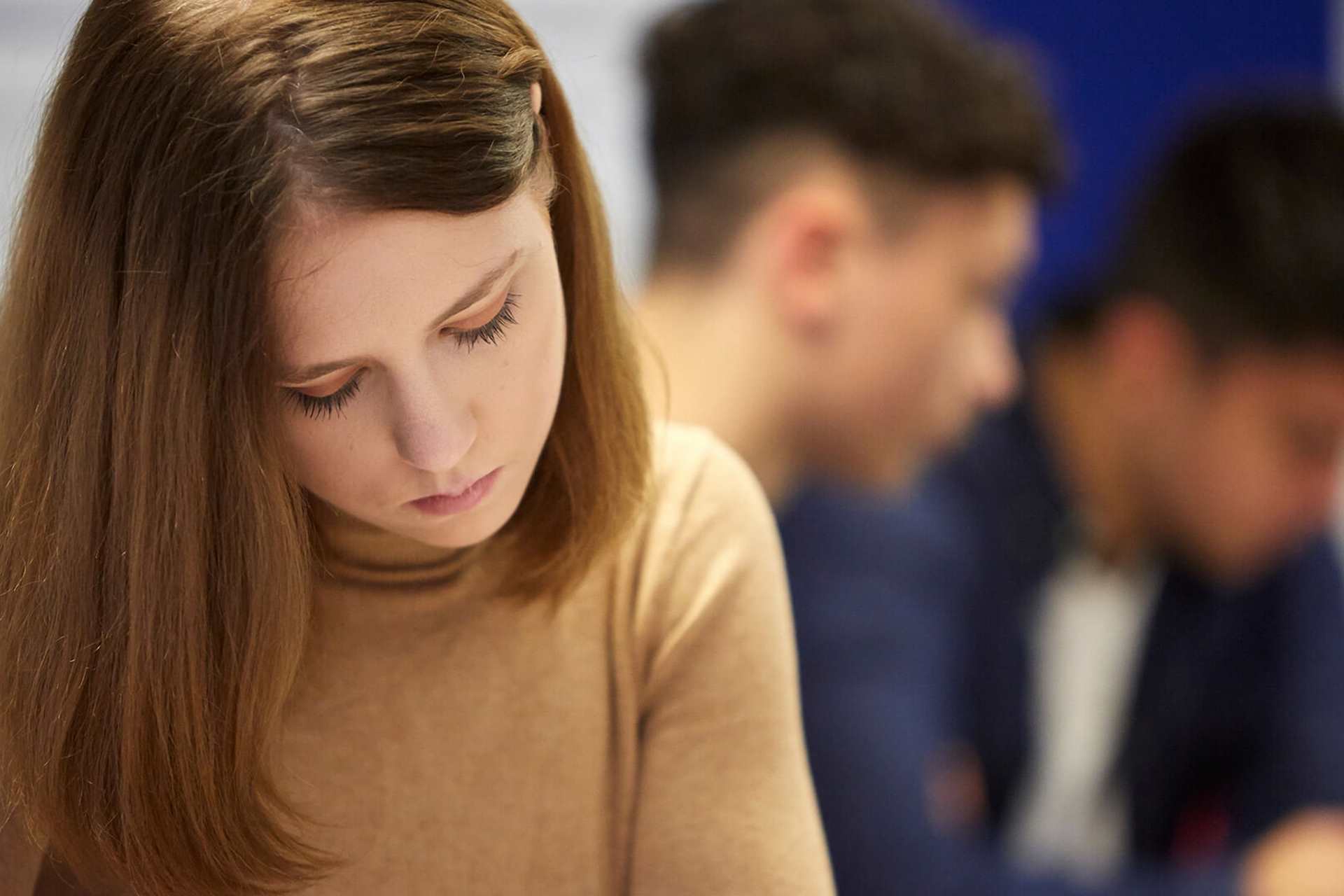 A young woman in a brown jumper looking down with a sad expression with two young men in the background.