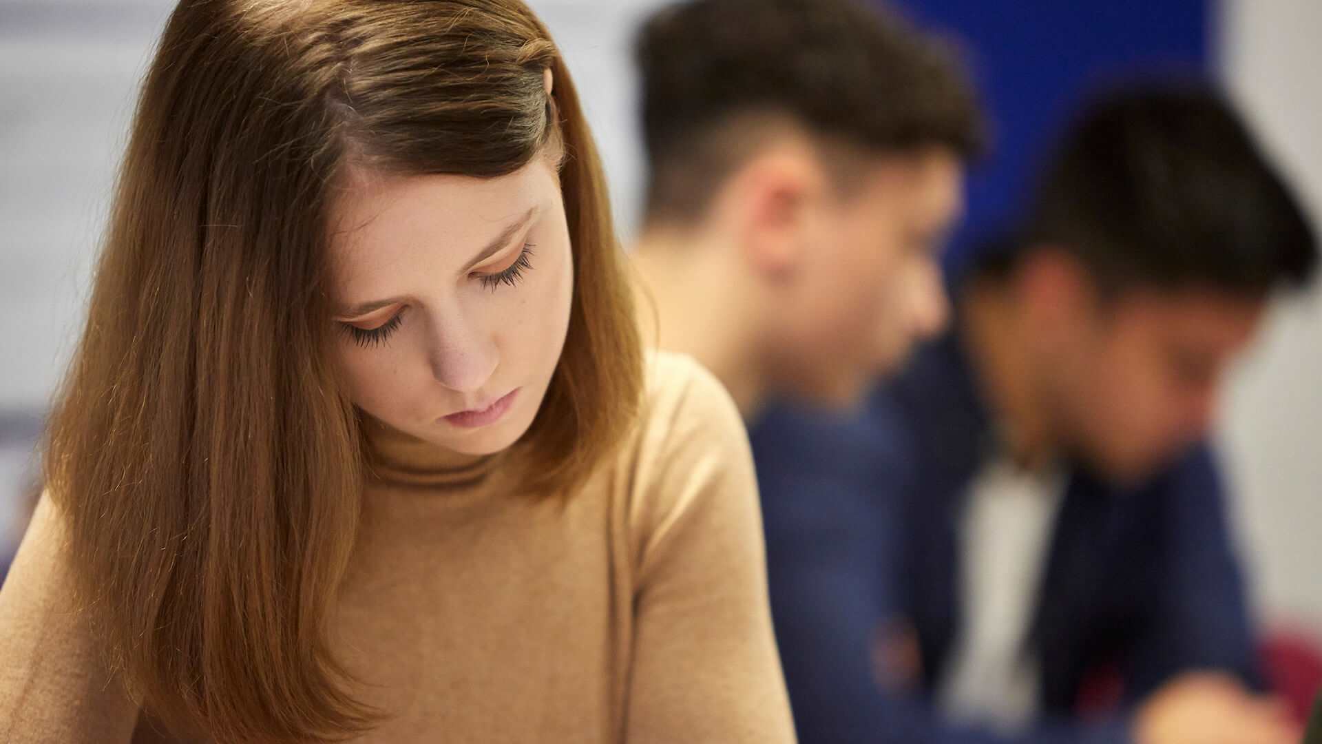 A young woman in a brown jumper looking down with a sad expression with two young men in the background.