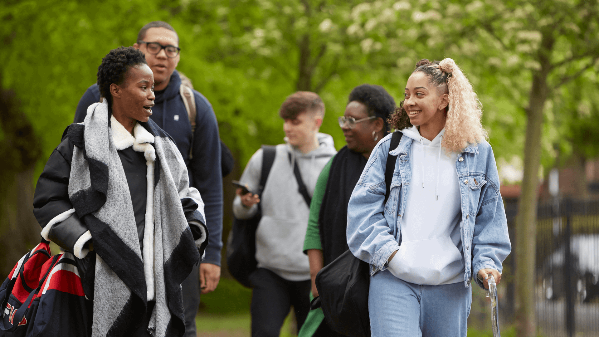 wide-shot-of-five-young-people-talking-and-smiling-while-walking-in-a-park-with-trees