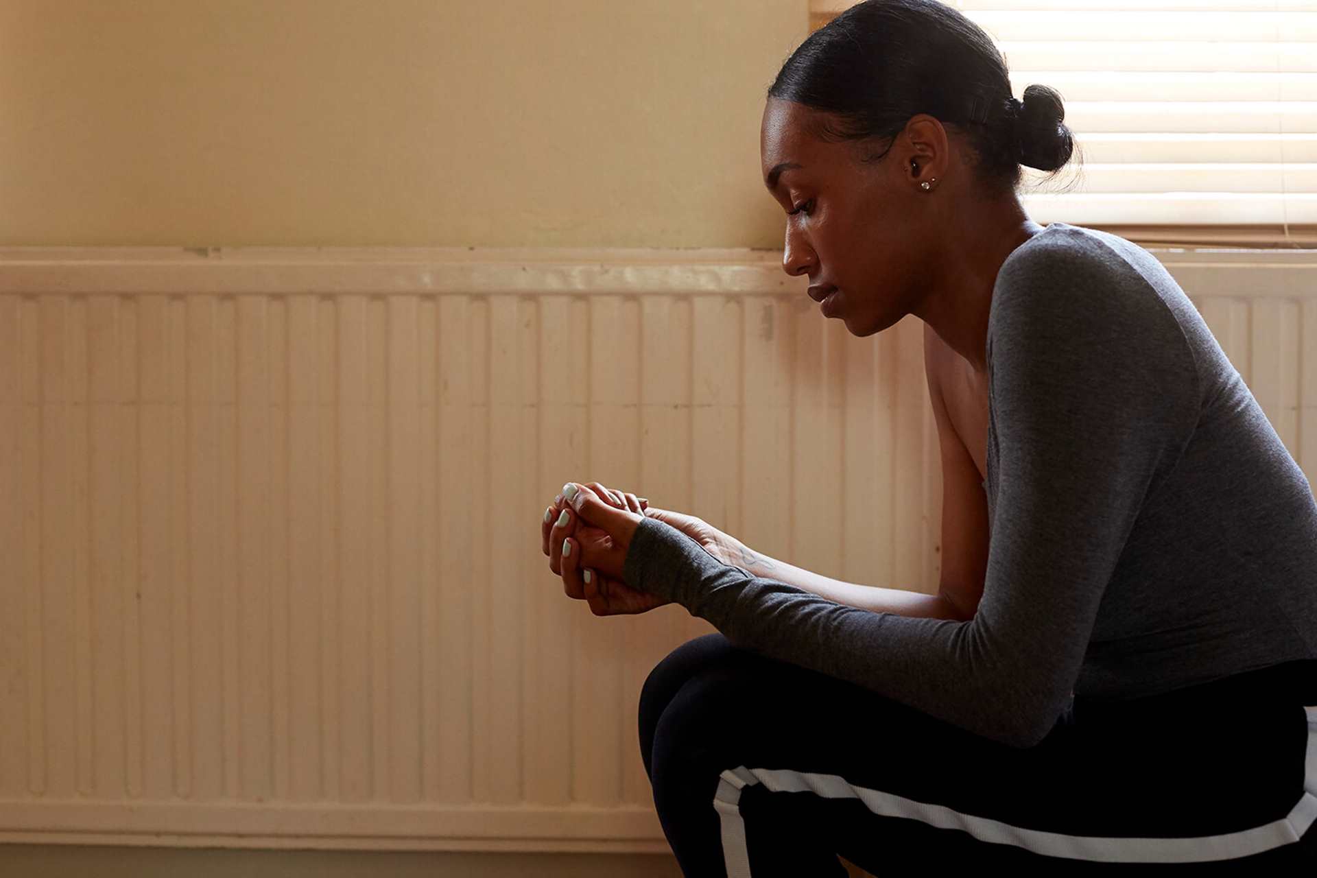 A girl sitting on a sofa in a living room beside a radiator. She is hunched over and looking down at her hands clasped together.