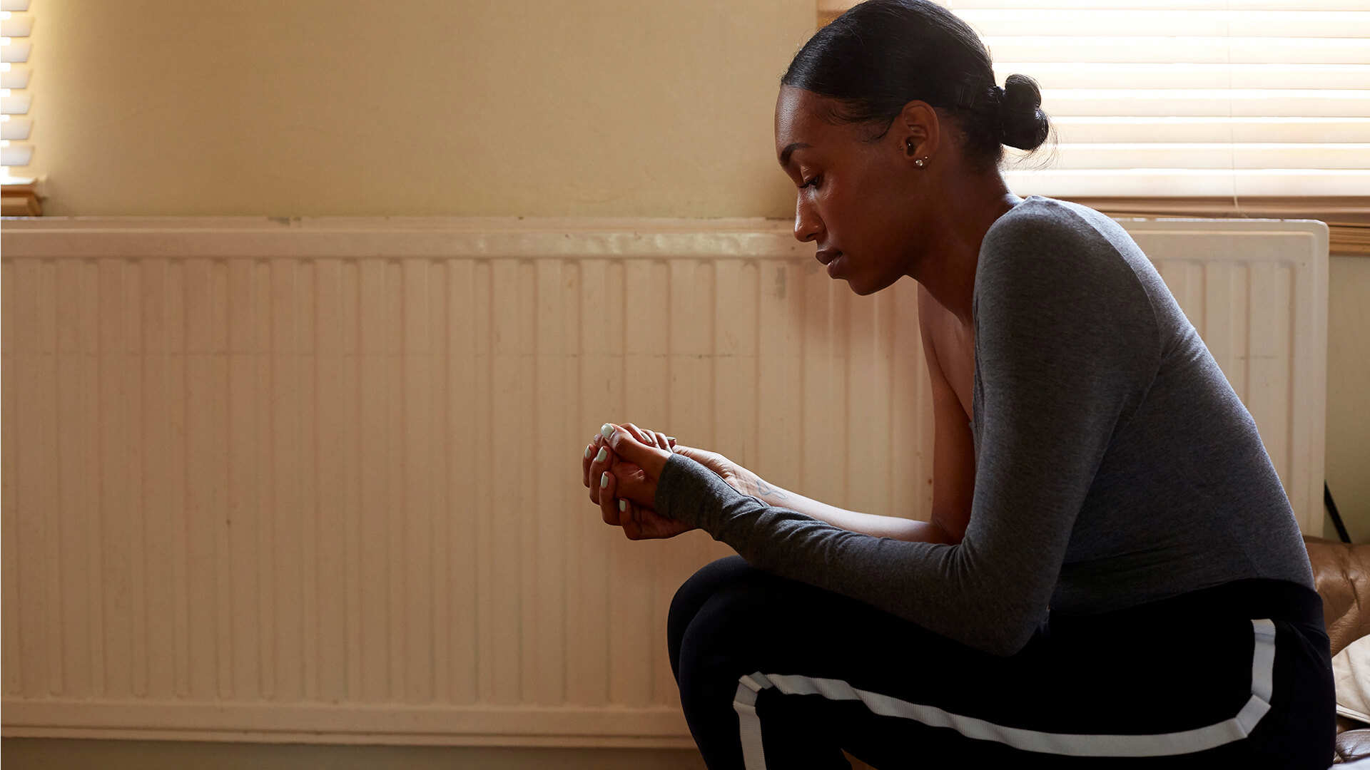 A girl sitting on a sofa in a living room beside a radiator. She is hunched over and looking down at her hands clasped together.