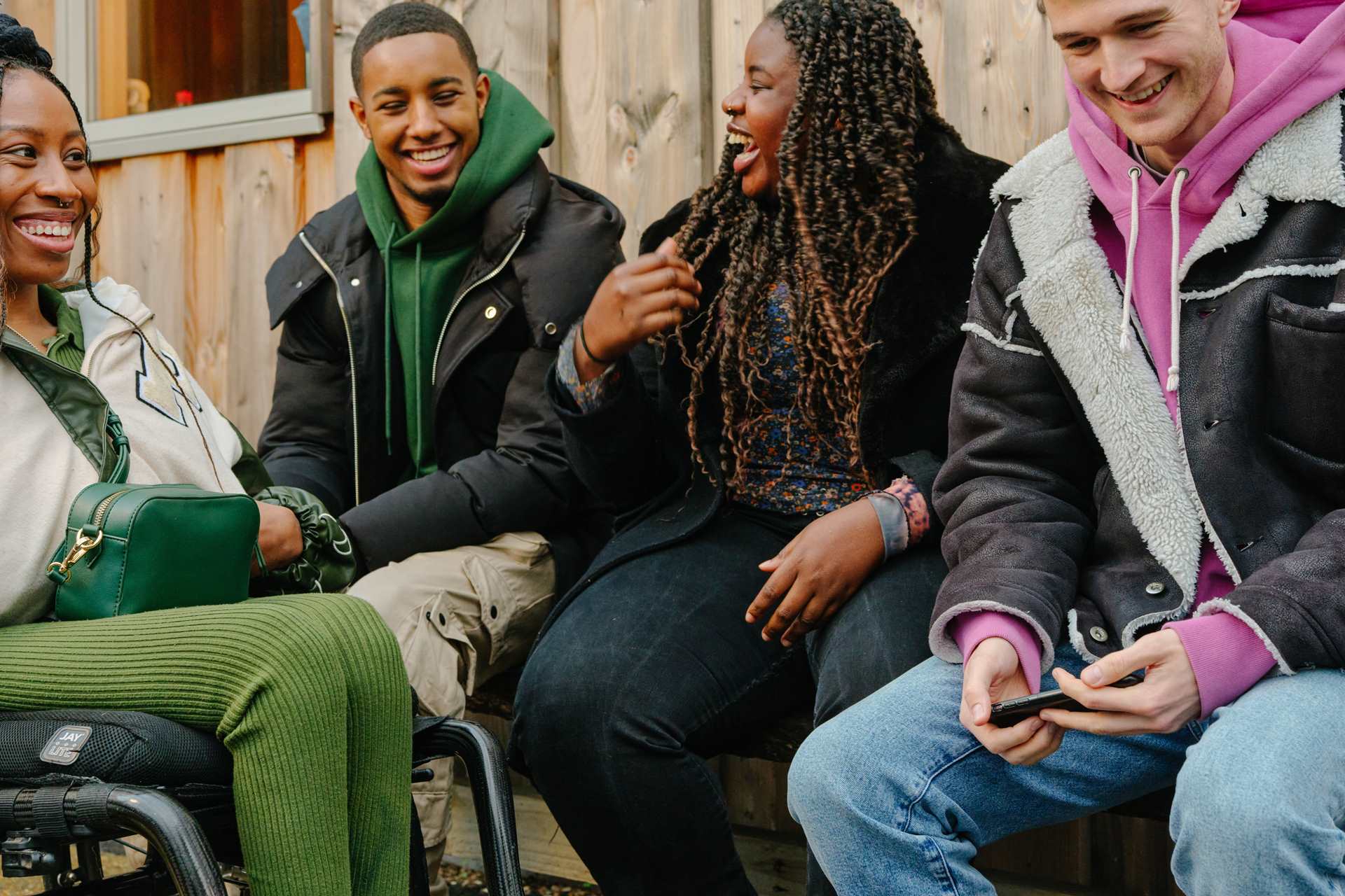 A group of young people laughing together outside on a bench. Group includes two Black young women (one in a wheelchair), one Black young man, and a white young man.