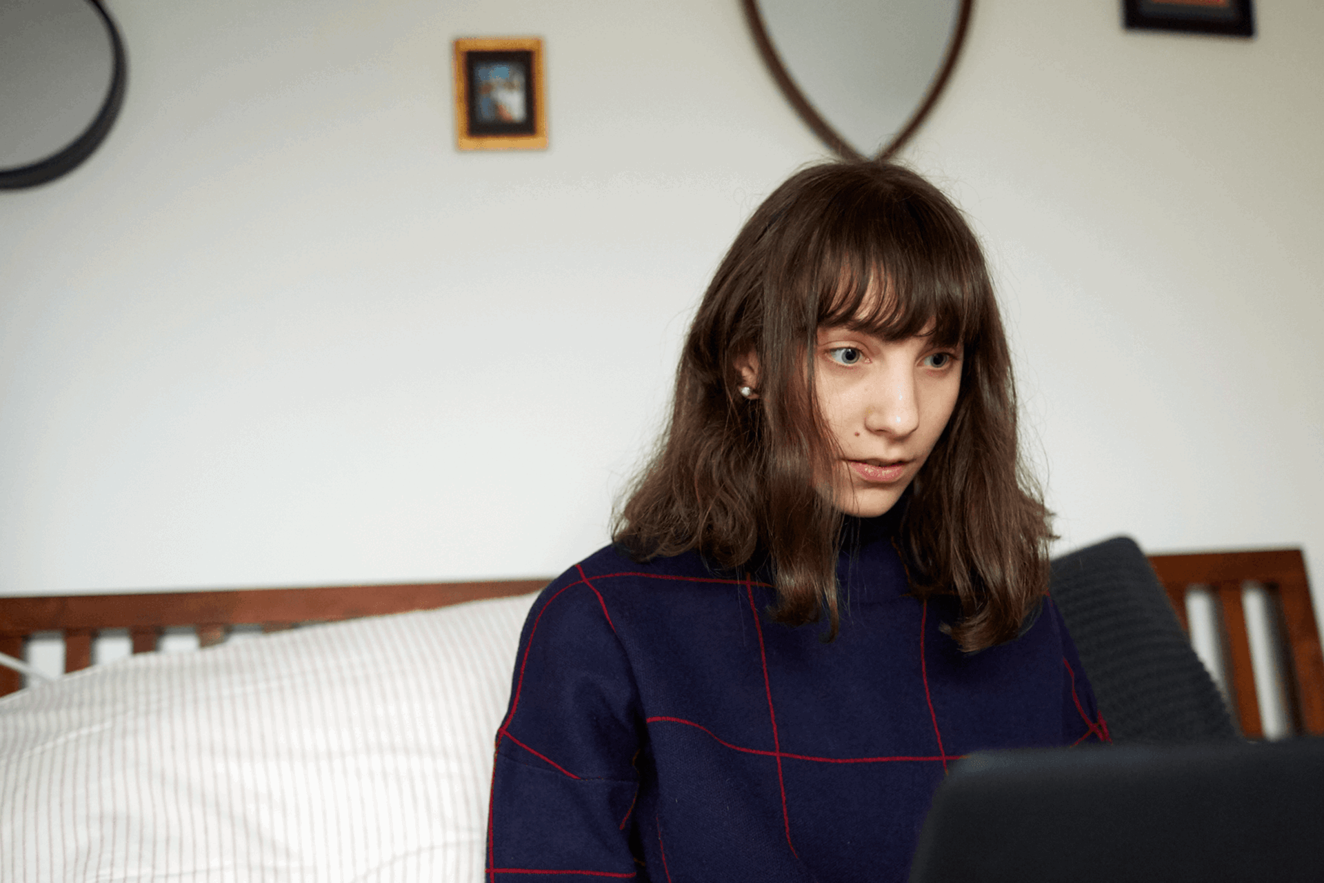 A girl sits on the end of her bed while looking at her laptop.