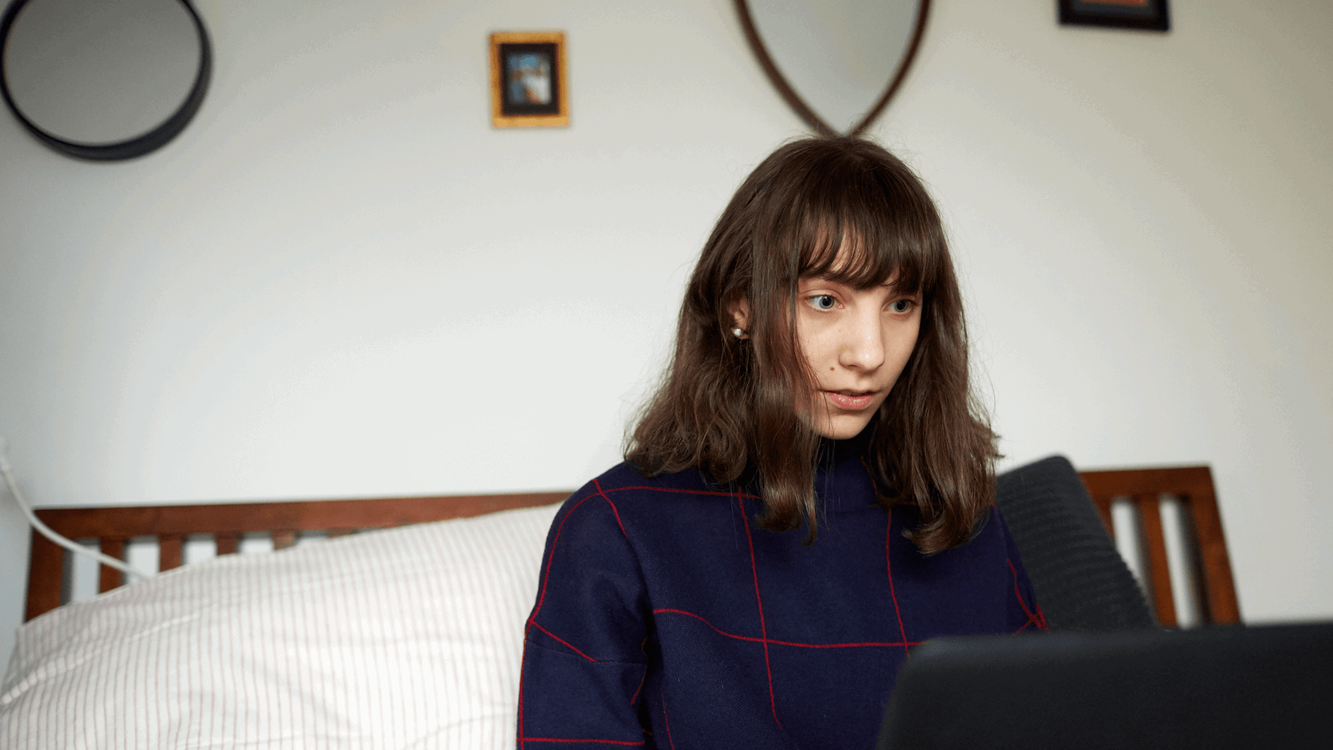 A girl sits on the end of her bed while looking at her laptop.