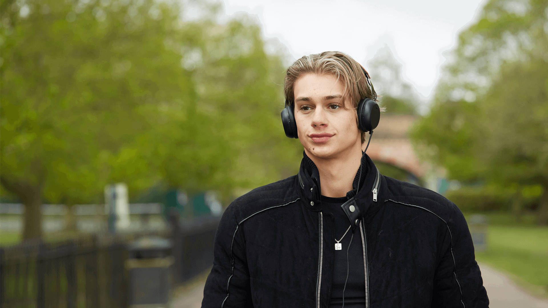 a boy wearing black jacket and with headphones on while walking on a tree lined street