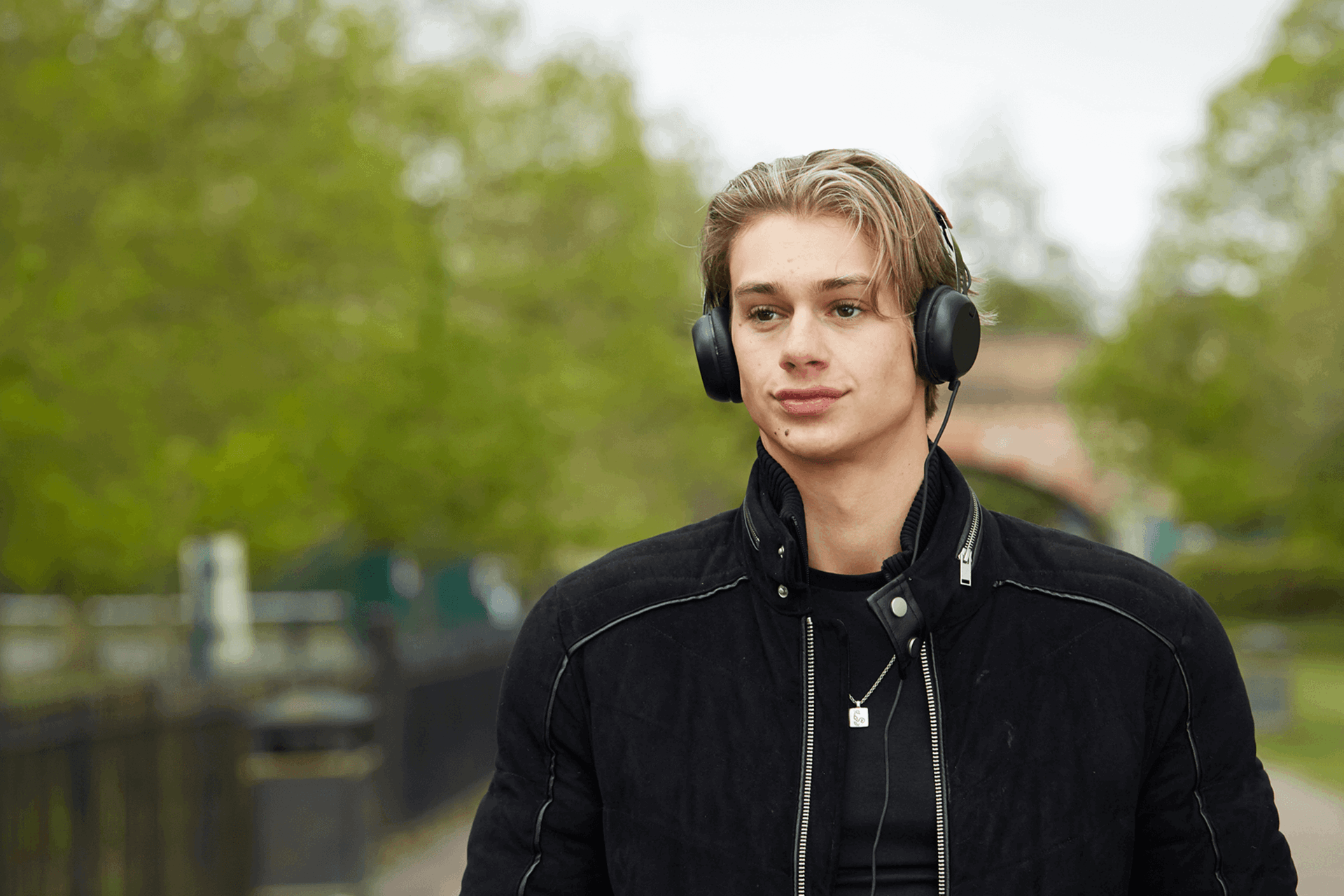 a boy wearing black jacket and with headphones on while walking on a tree lined street